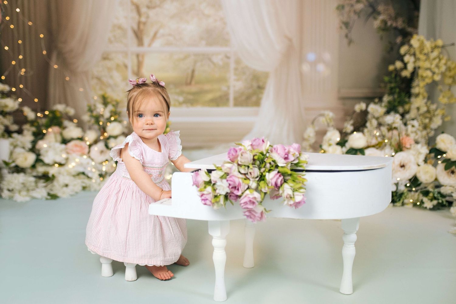 A toddler girl in a pink dress stands by a white mini grand piano adorned with flowers in a floral decorated studio.