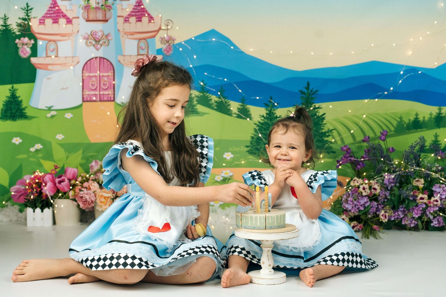Two girls in matching blue Alice in Wonderland dresses celebrate a birthday with cake against a fairy tale castle backdrop.
