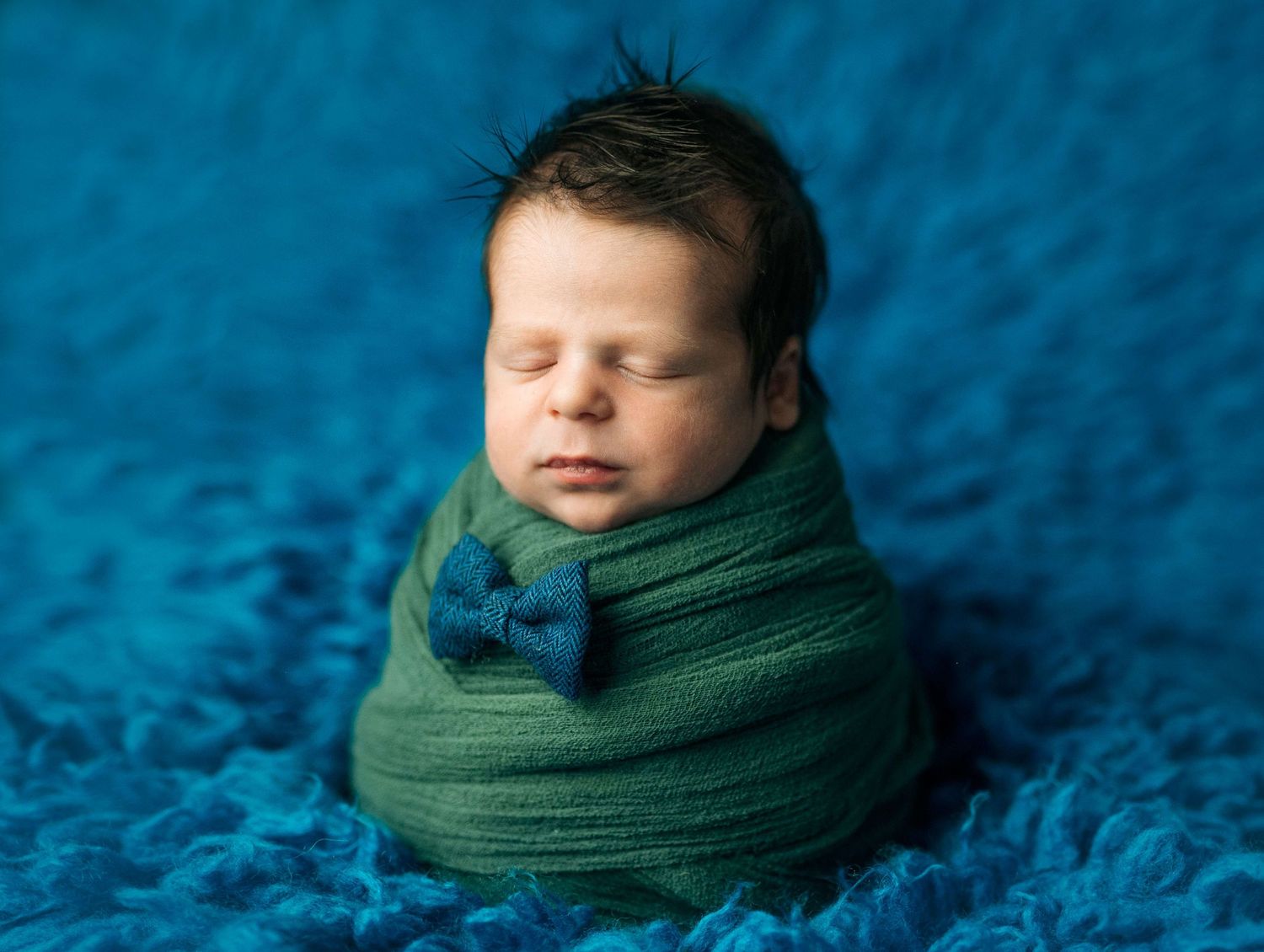 Newborn baby swaddled in green wrap with bow tie, sleeping peacefully on blue textured blanket.