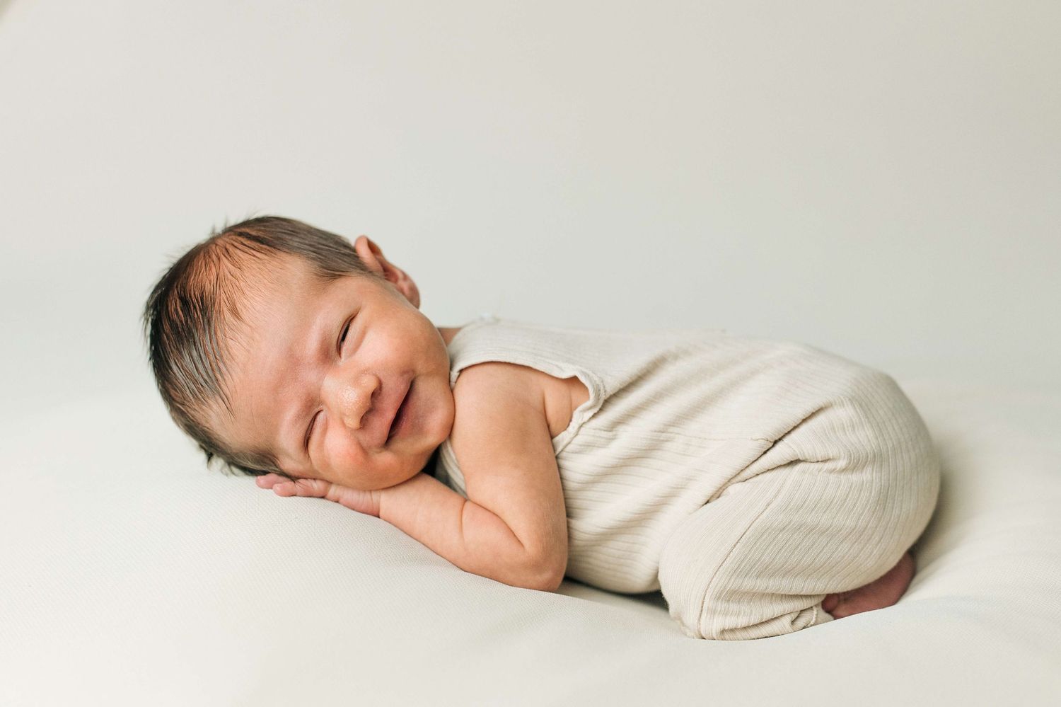 Smiling newborn baby in cream romper lying on white blanket during professional newborn photography session.