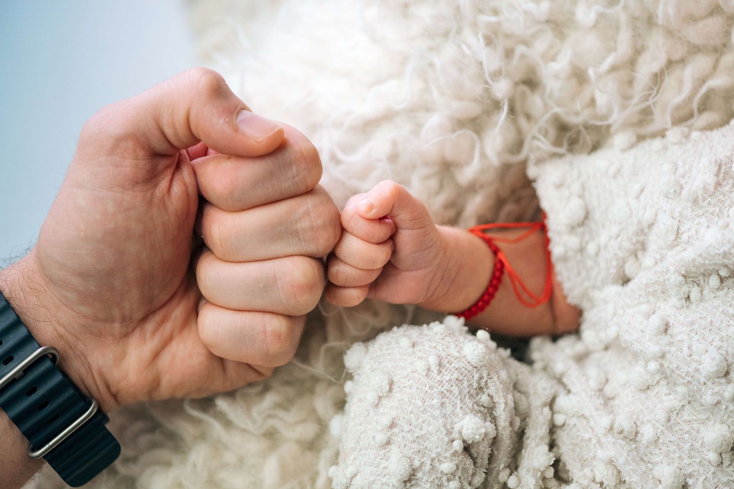 Adult and newborn baby doing a fist bump, with baby wrapped in white fluffy blanket wearing a red bracelet.