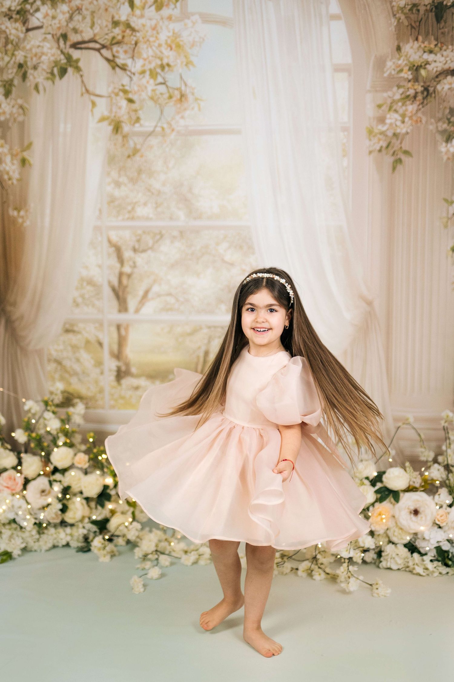 Young girl in pink dress twirling barefoot among floral backdrop with white curtains and spring blossoms.