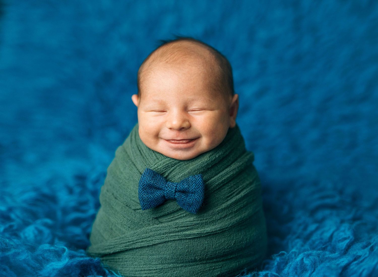Smiling newborn baby swaddled in green wrap with blue bow tie on blue fluffy background.