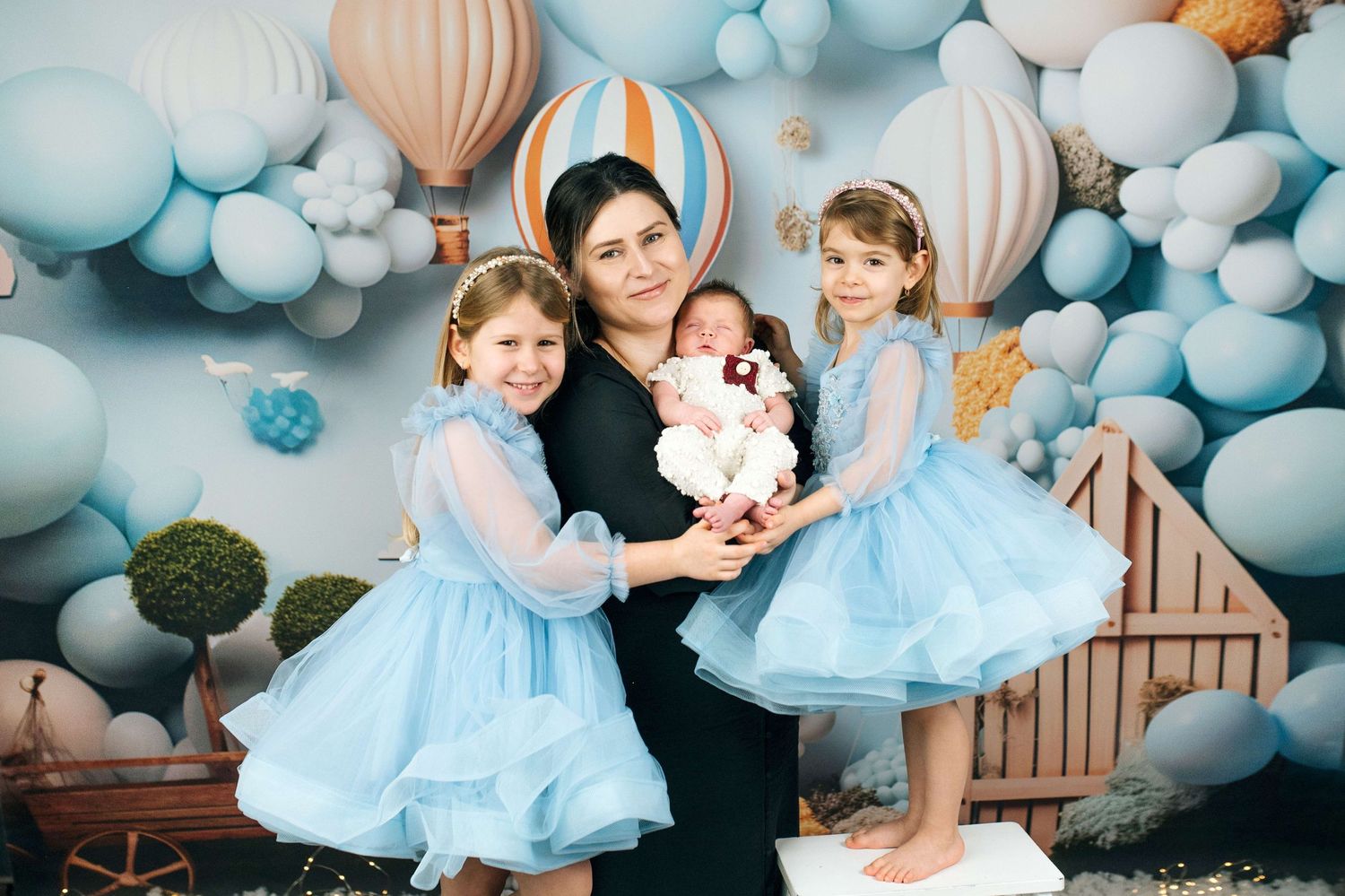 Woman in black dress holds newborn baby flanked by two girls in blue tutus, with balloon backdrop.