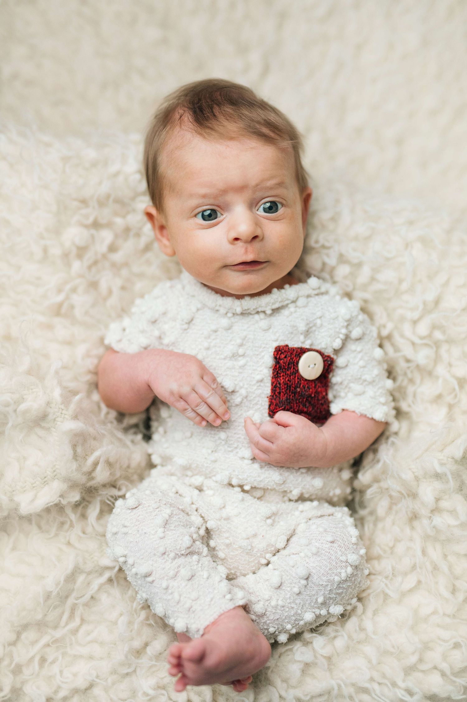 Adorable baby in white knit outfit with red pocket detail lying on fluffy cream blanket, looking up with bright blue eyes.