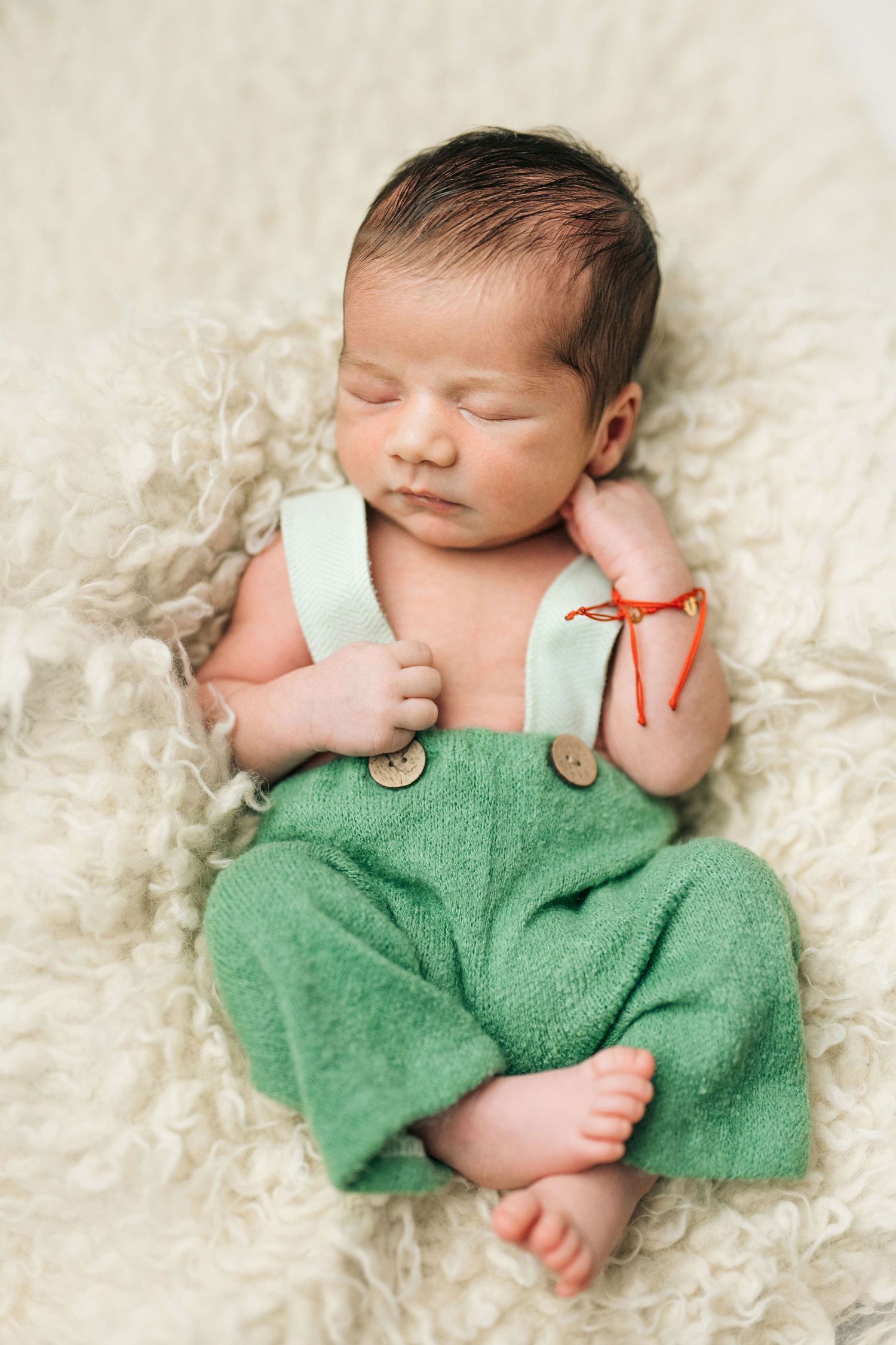 Sleeping newborn baby wearing green suspender pants on a fluffy cream blanket.