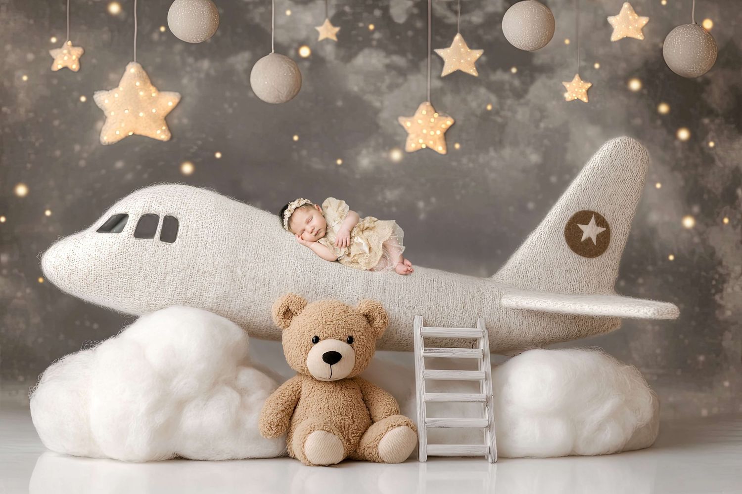 Newborn baby sleeping on a plush toy airplane with a teddy bear, stars, and clouds in a dreamy nursery photo.