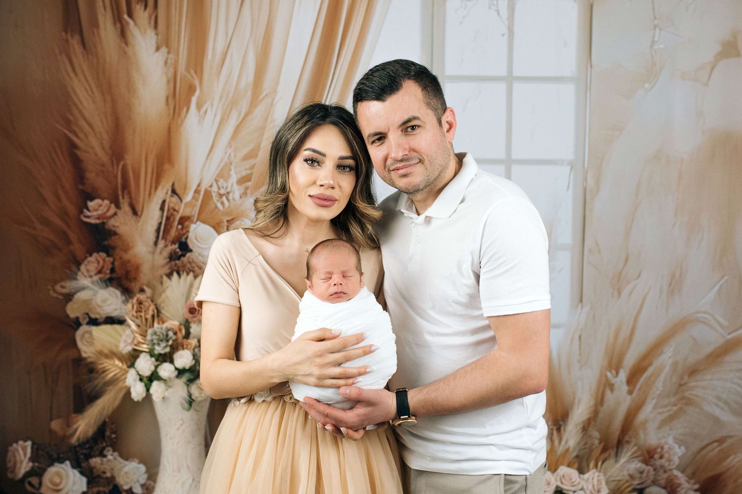 Happy couple holding their newborn baby in a boho-styled photo studio with floral decorations.