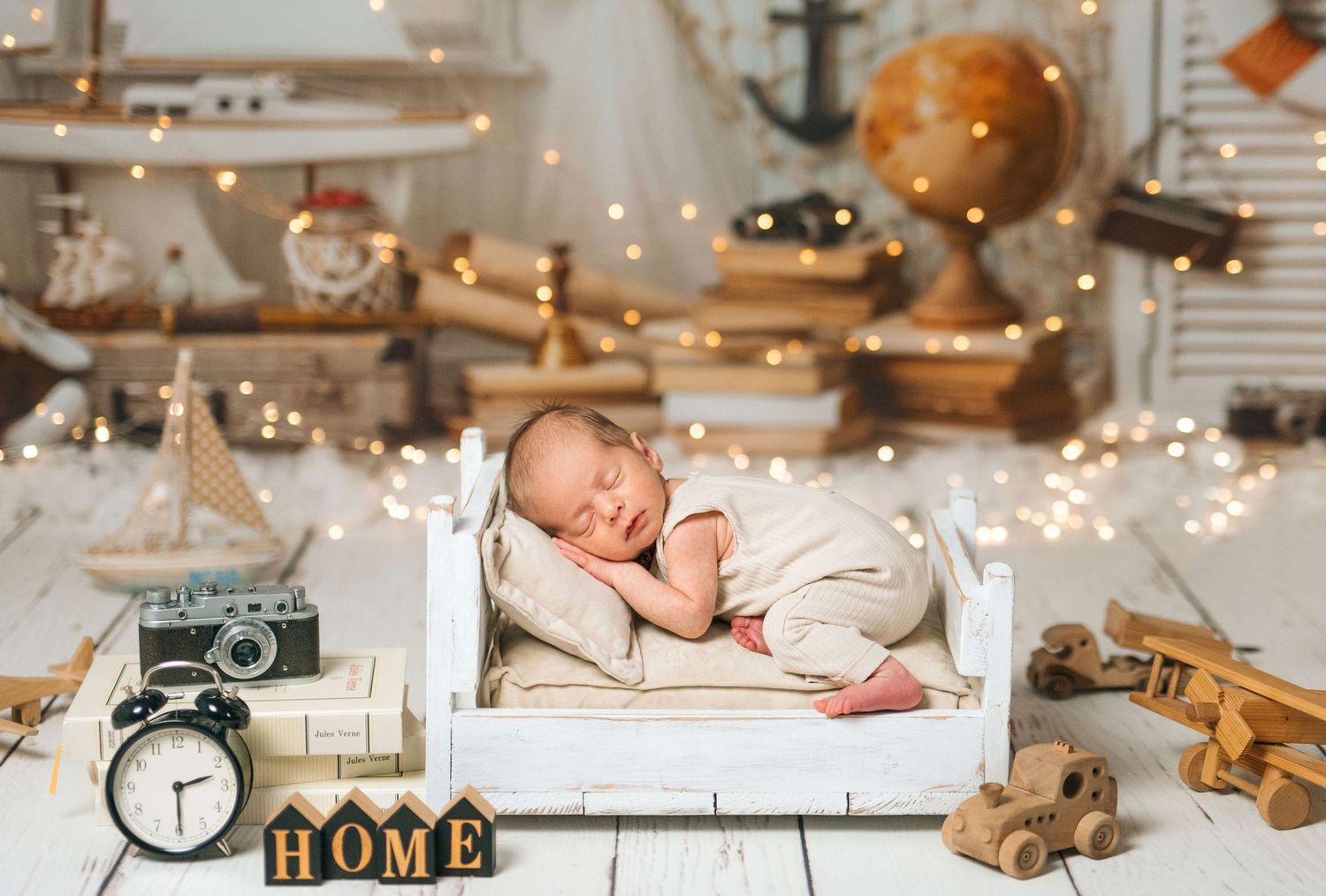 Newborn baby sleeping peacefully on a tiny white bed surrounded by vintage toys, a globe, and warm fairy lights.