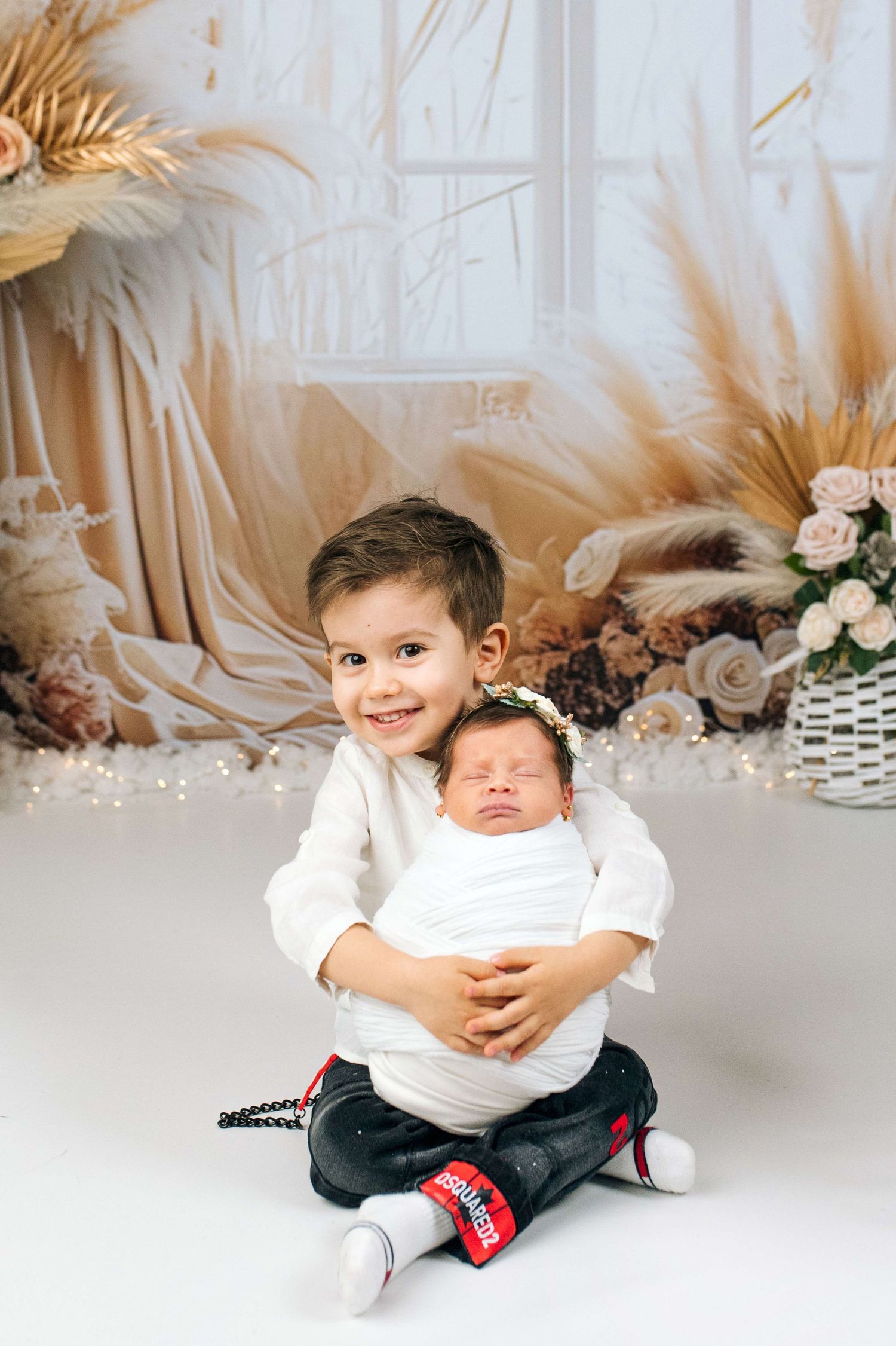 Young boy smiling while holding swaddled newborn baby sibling in a decorated studio photo session.
