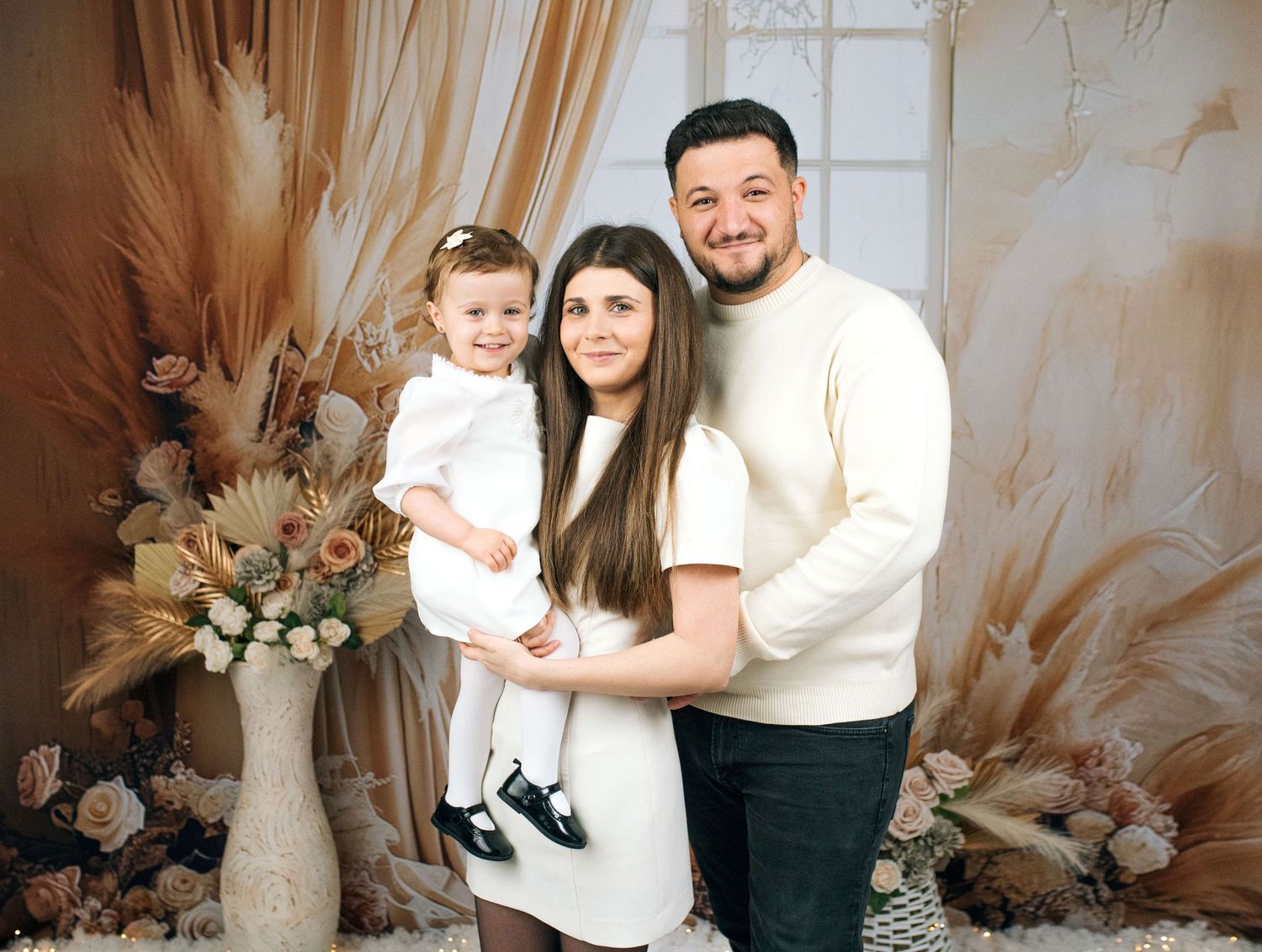 Happy family of three posing in a boho-styled photo studio with floral decor, wearing white outfits.