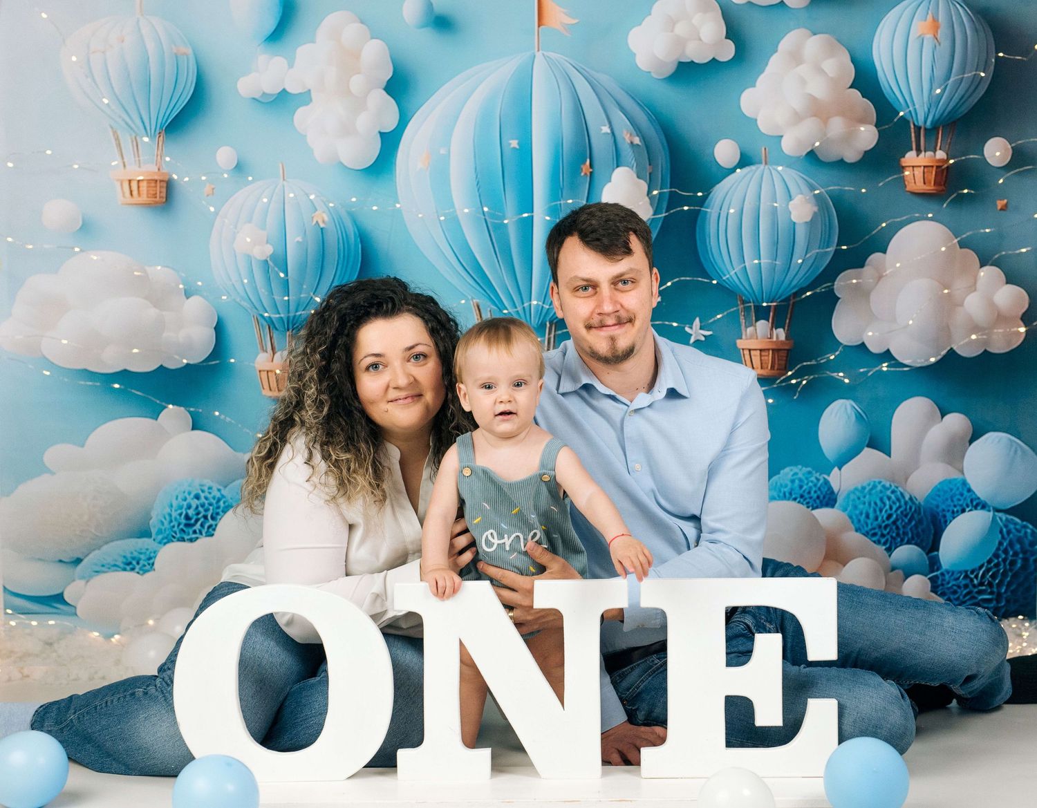 Family of three celebrating baby’s first birthday with blue hot air balloon backdrop and ONE sign.