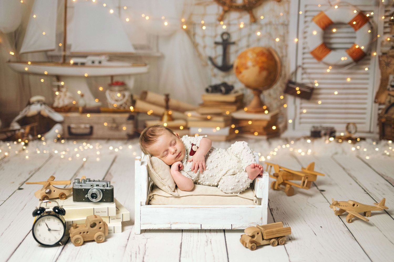 Newborn baby sleeping peacefully in a tiny white bed surrounded by vintage toys and warm fairy lights.