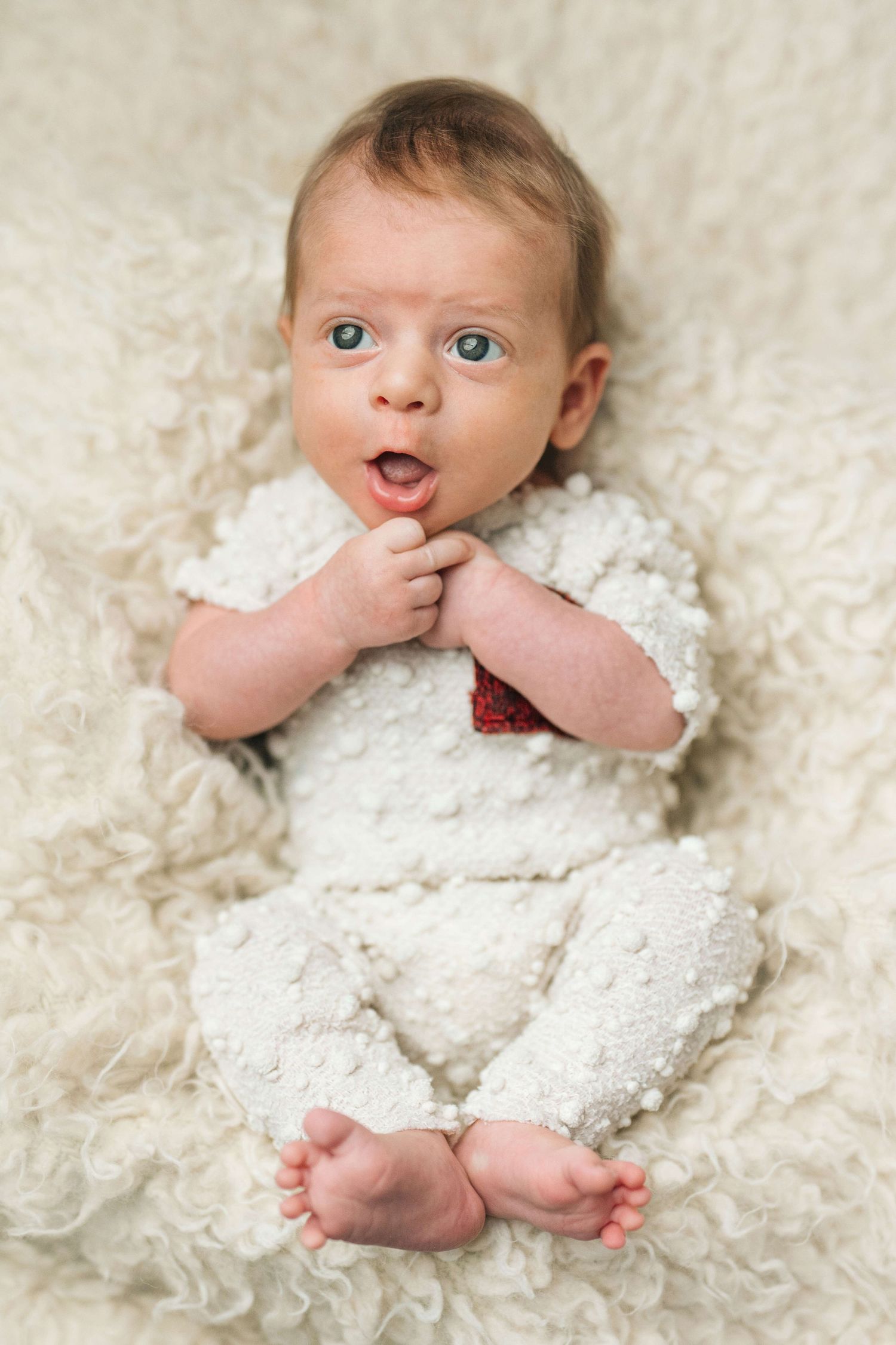 Adorable blue-eyed baby in white knit outfit lying on fluffy cream blanket with mouth open in surprise.
