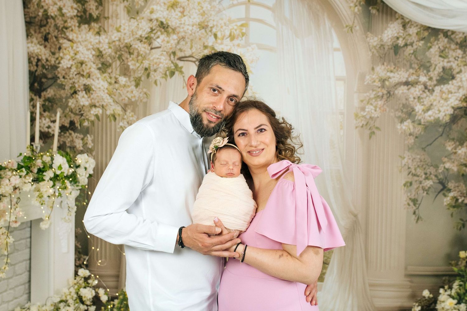 Family portrait with parents holding newborn baby in floral studio setting.