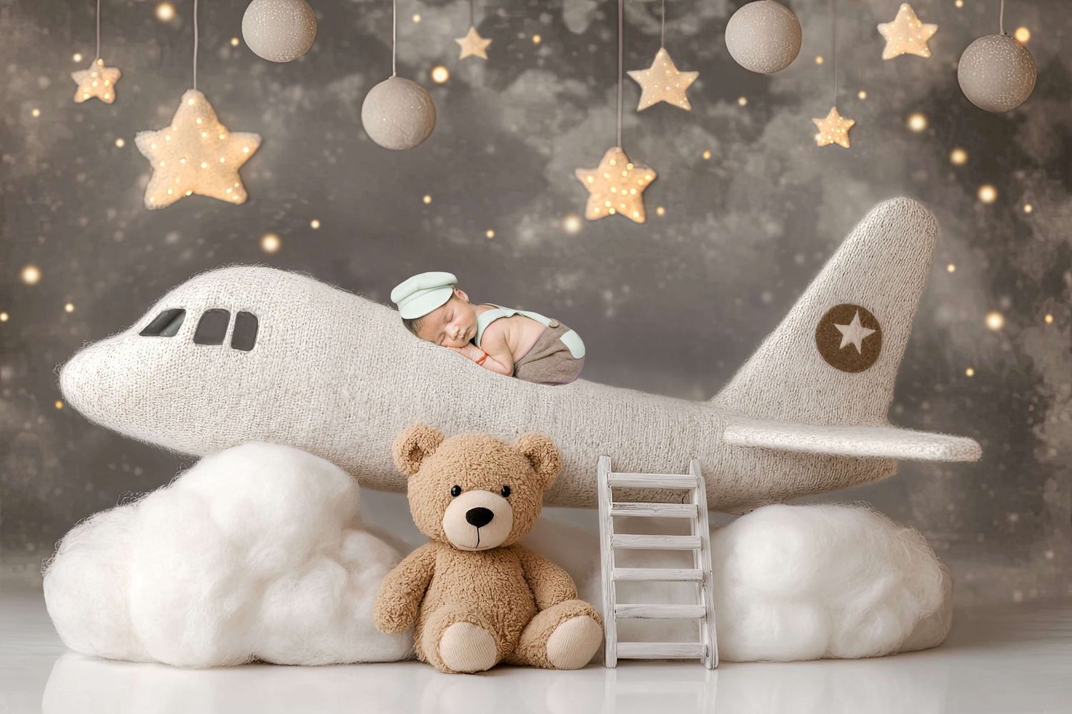 Newborn baby sleeping on a plush toy airplane with a teddy bear, stars, and clouds in a dreamy nursery photo.