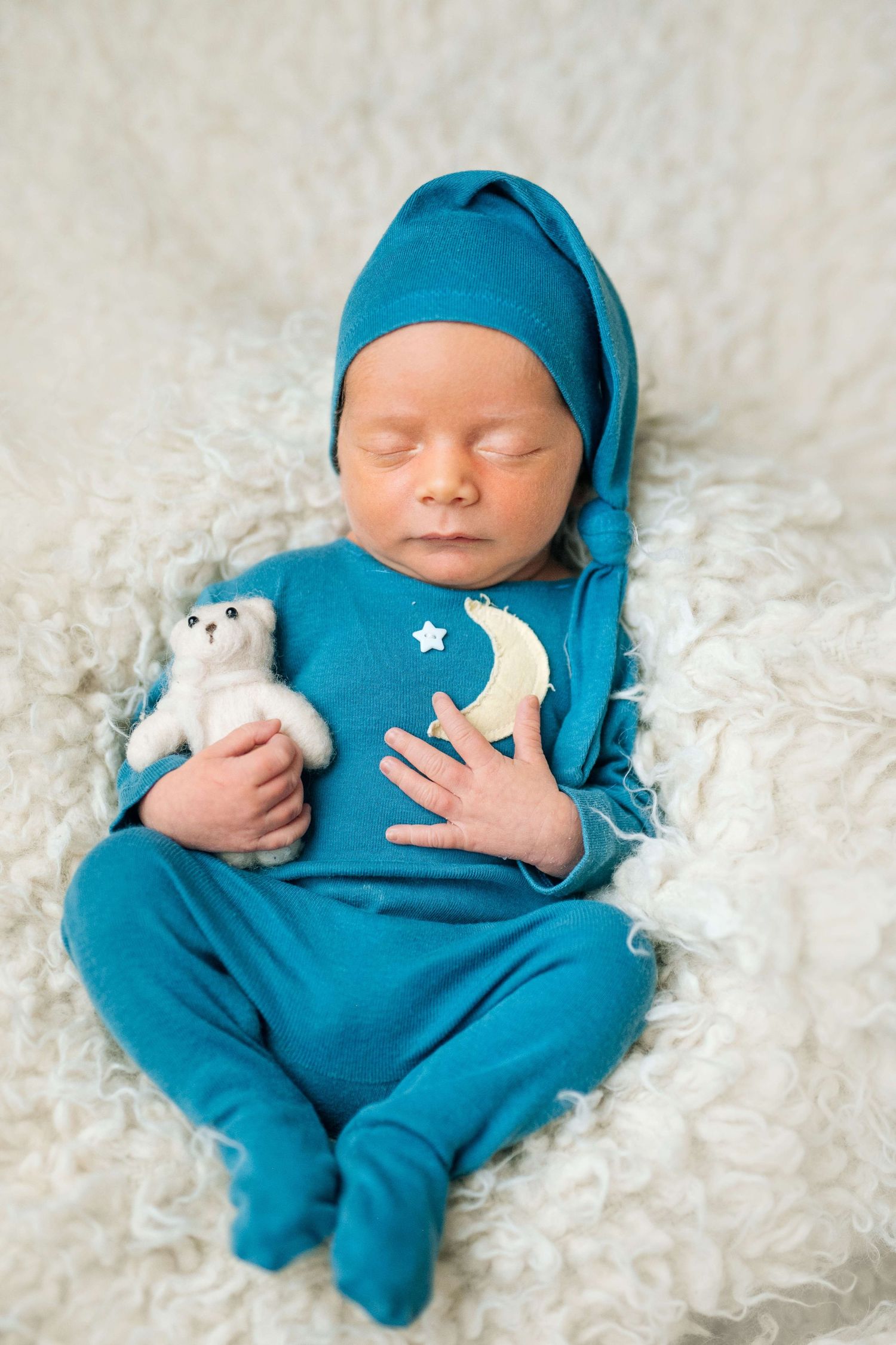 Newborn baby sleeping in teal outfit and hat, holding a small white teddy bear on a fluffy white blanket.