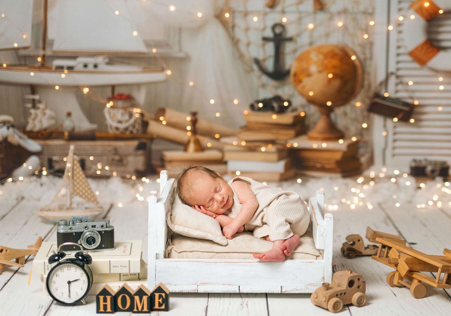 Newborn baby sleeping peacefully in a tiny white bed surrounded by vintage toys, wooden decor, and warm fairy lights.