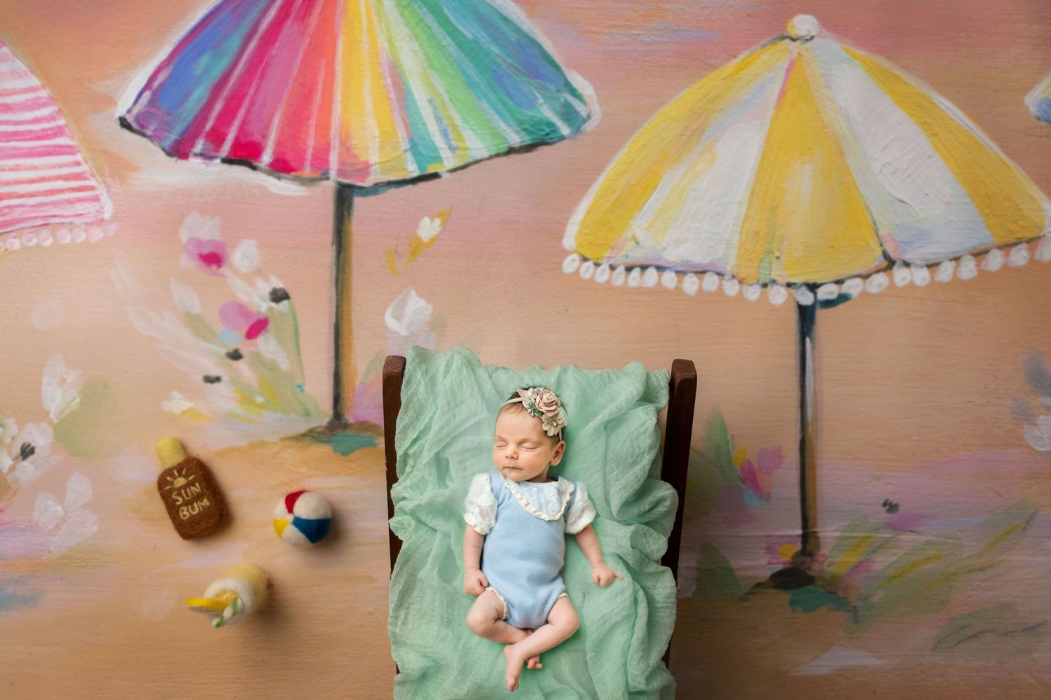 Newborn baby sleeping on a green blanket lounge chair surrounded by colorful beach umbrella painted backdrop.