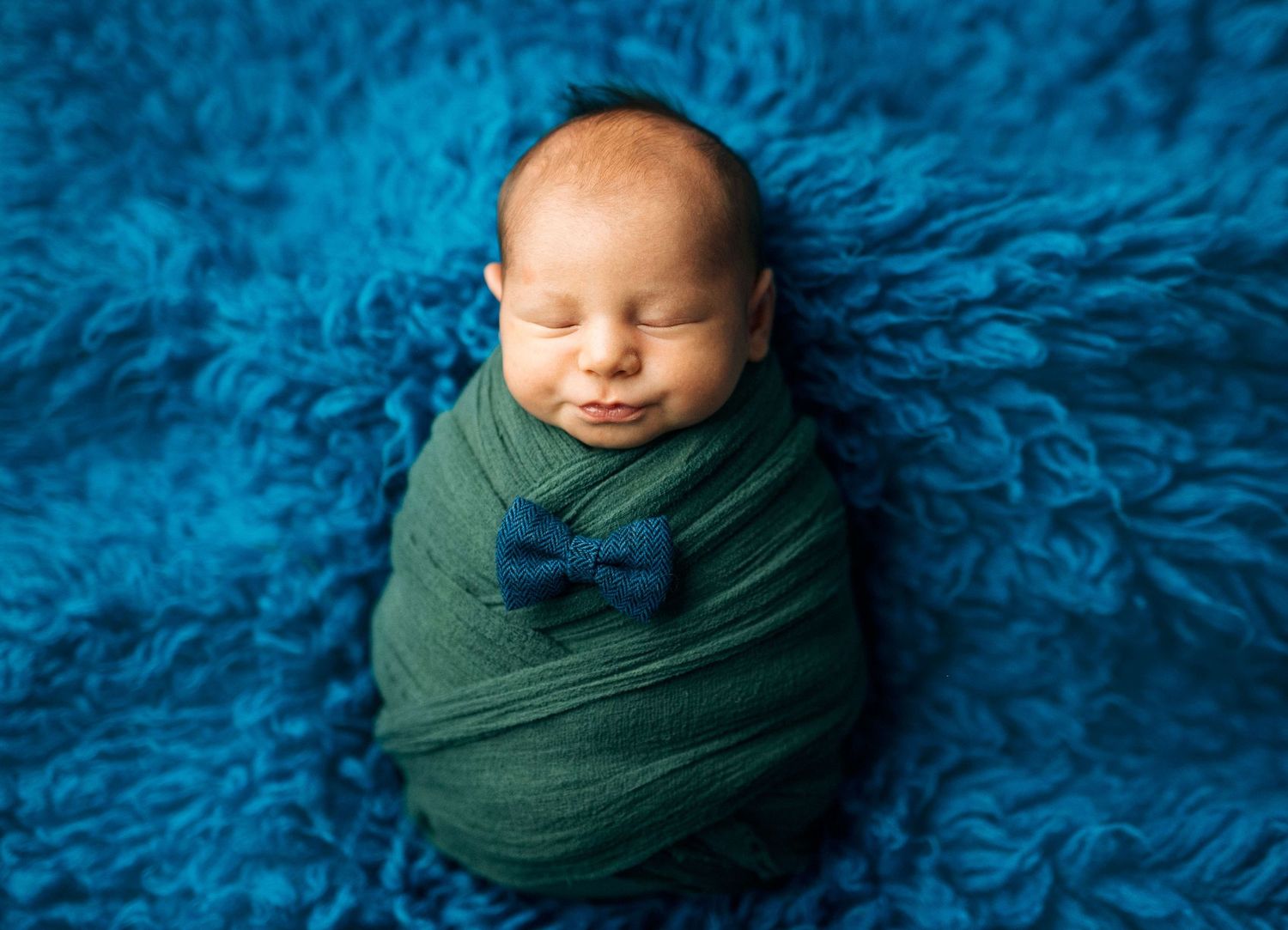 Newborn baby swaddled in green wrap with blue bow tie, sleeping on blue fluffy rug.