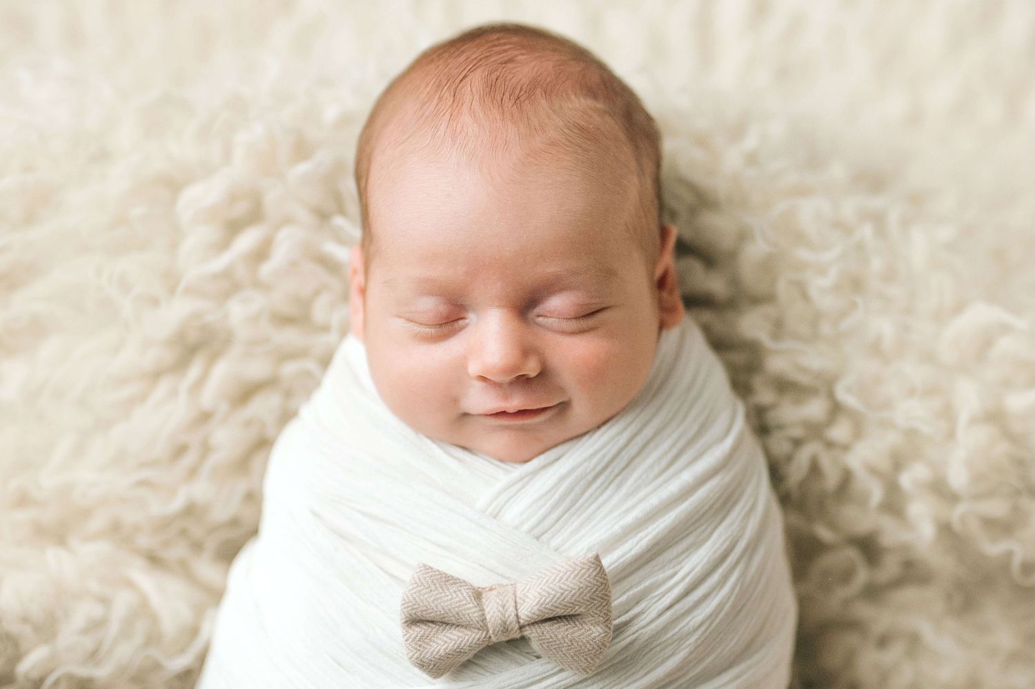 Smiling newborn baby swaddled in white wrap with a bow, lying on a fluffy cream blanket.