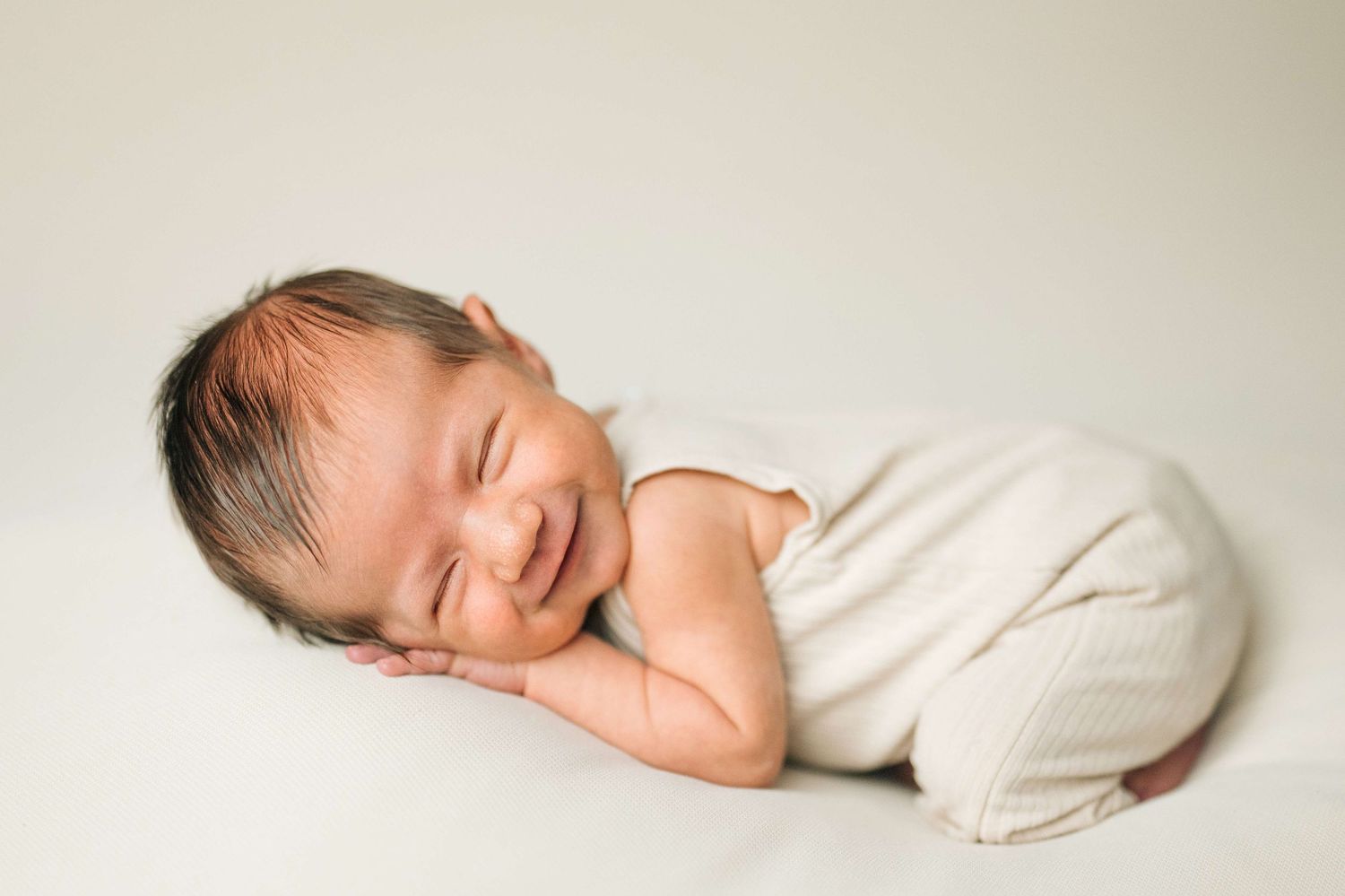 Smiling newborn baby wrapped in cream fabric, peacefully sleeping on a white background.