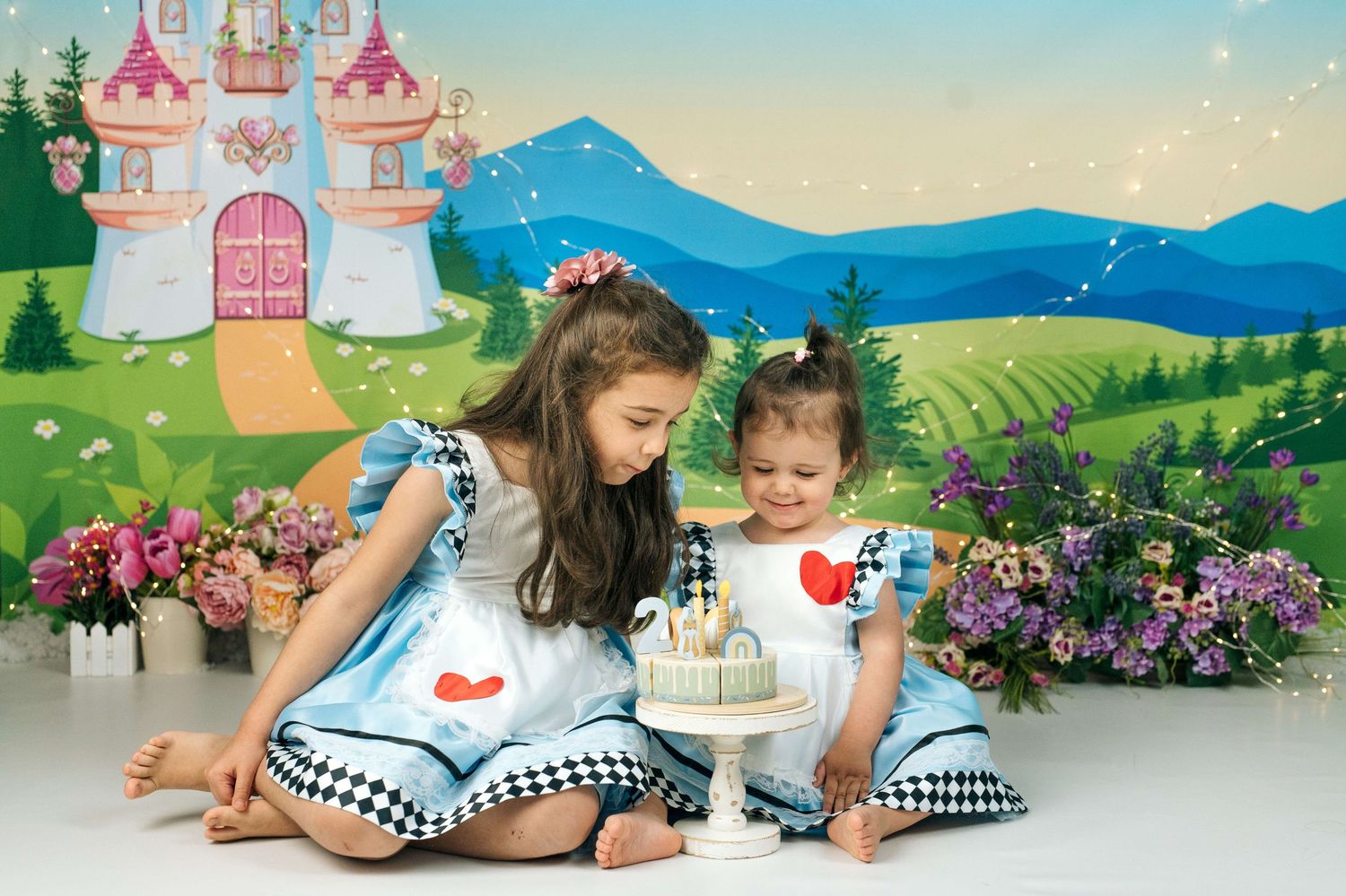 Two young girls in matching Alice in Wonderland dresses sit together examining a small cake against a fairy tale castle backdrop.