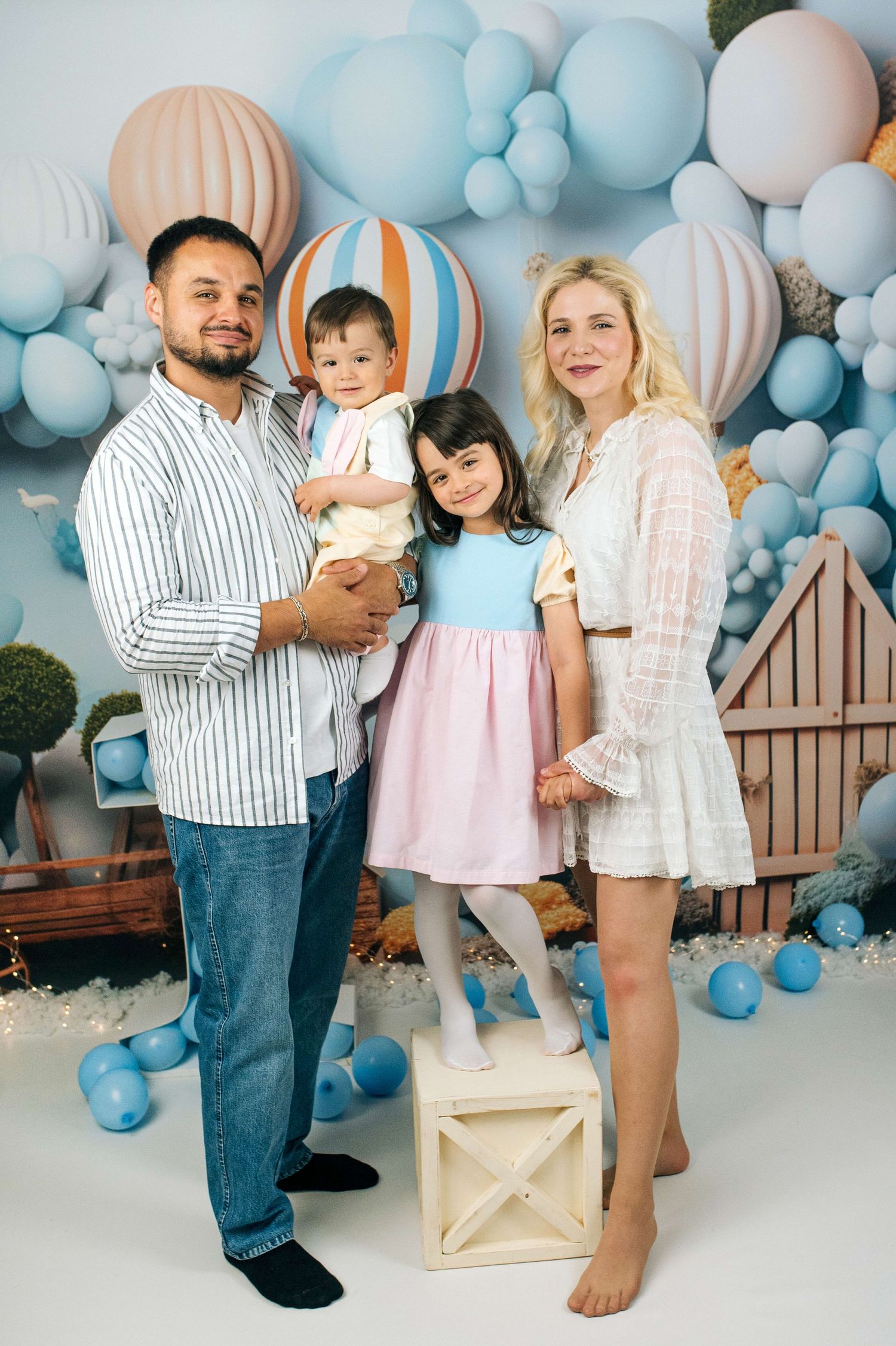 Happy family of four posing in a festive studio with blue balloon decorations and hot air balloon backdrop.