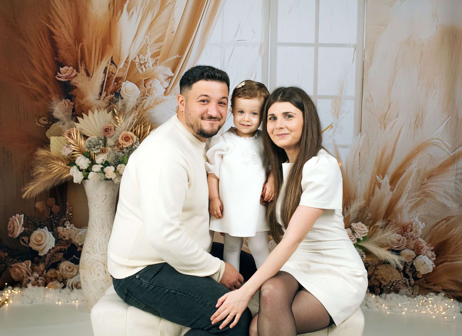 Family portrait of parents and young daughter in white outfits posed with boho floral decor backdrop.