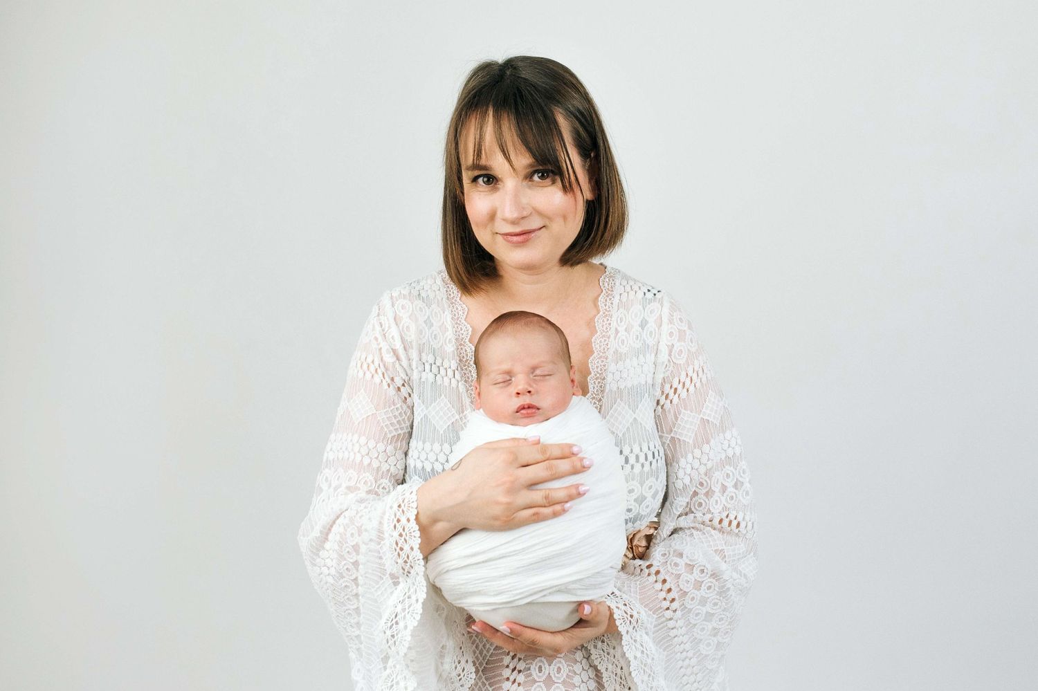 A young mother in a white lace dress tenderly holds her sleeping newborn baby wrapped in white cloth.