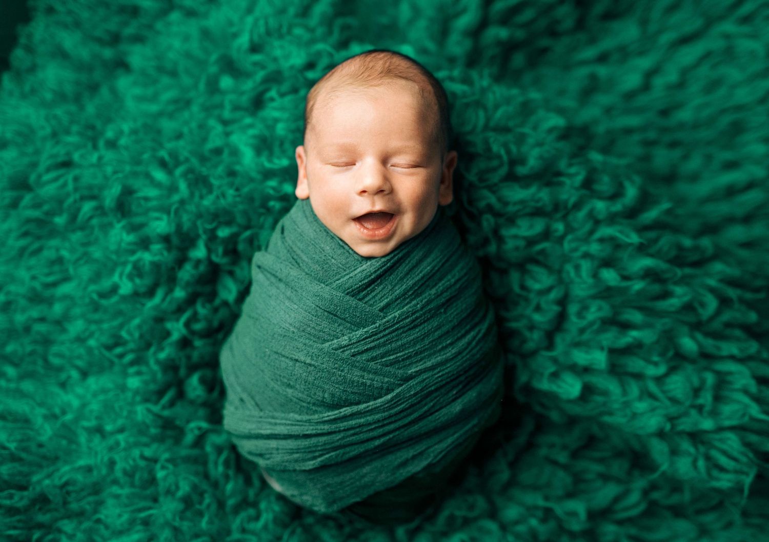 Smiling newborn baby swaddled in green wrap on fluffy emerald blanket.