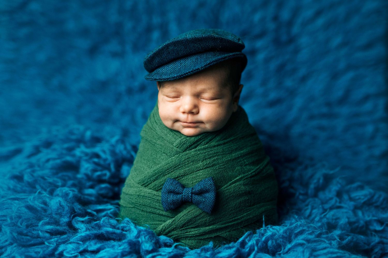 Newborn baby wrapped in green swaddle wearing flat cap and bow tie, sleeping on blue fluffy blanket.