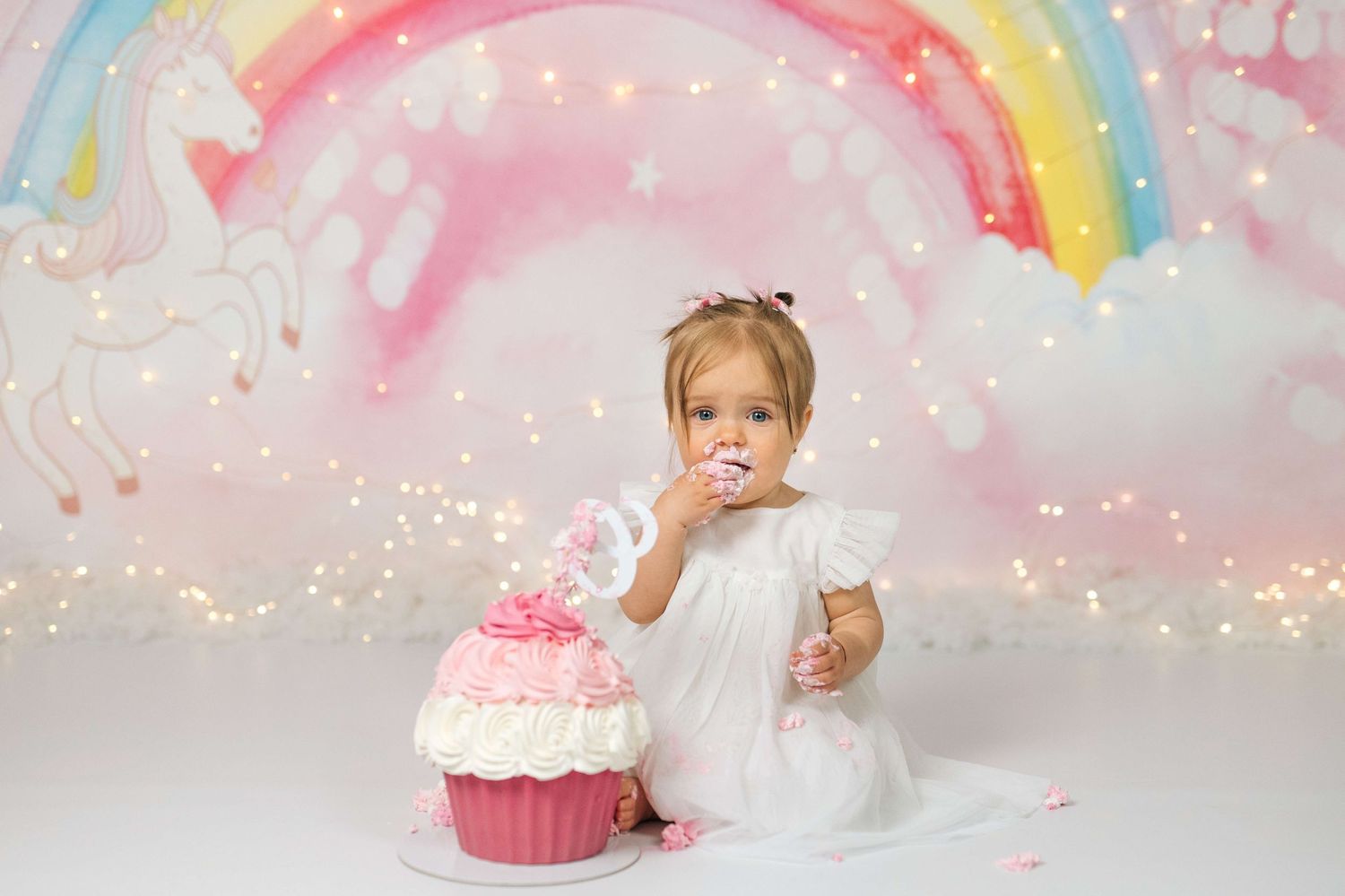 Toddler girl in white dress eating cake at unicorn rainbow birthday smash cake photo session.