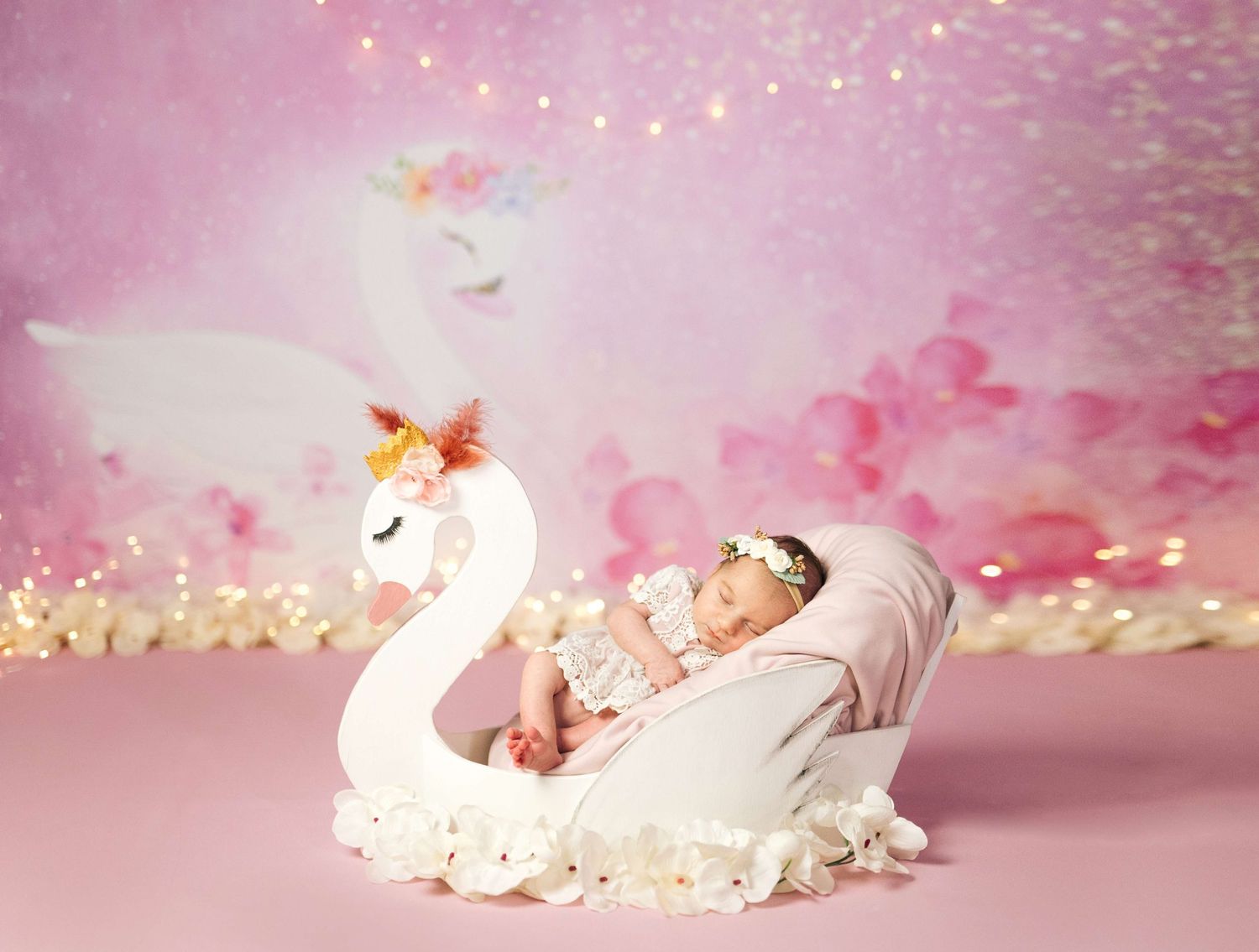 Newborn baby sleeping in a white swan-shaped prop on a pink glittery background for a fairy tale photo shoot.