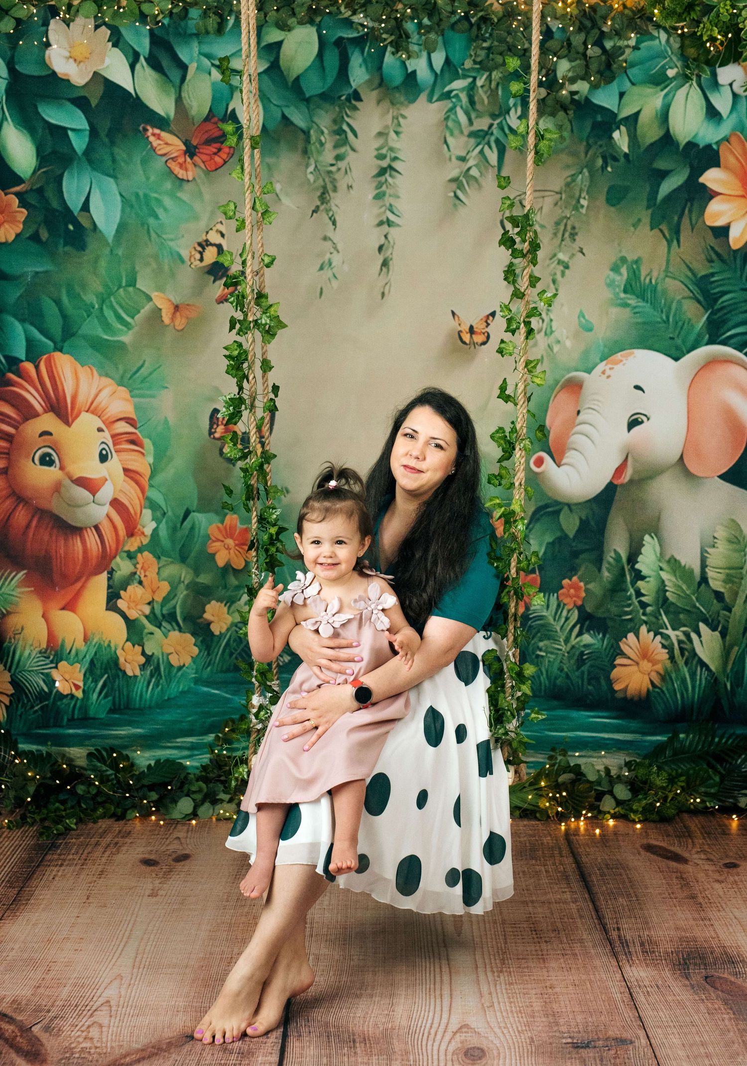 A woman and toddler girl sit on a jungle-themed swing surrounded by cartoon lions and elephants on a colorful backdrop.