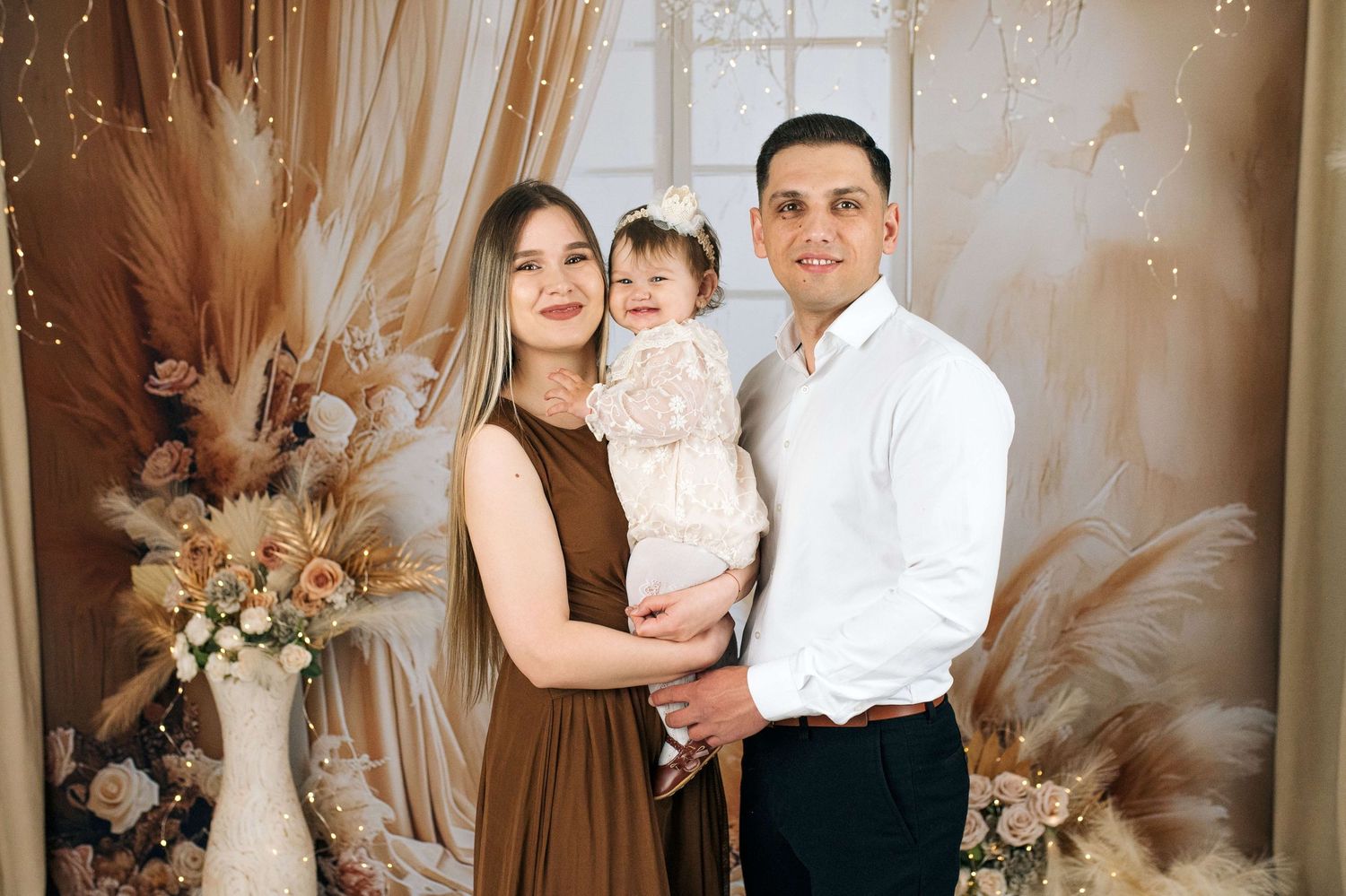 A young family poses together in a boho-styled photo shoot with warm brown tones, florals, and pampas grass decor.