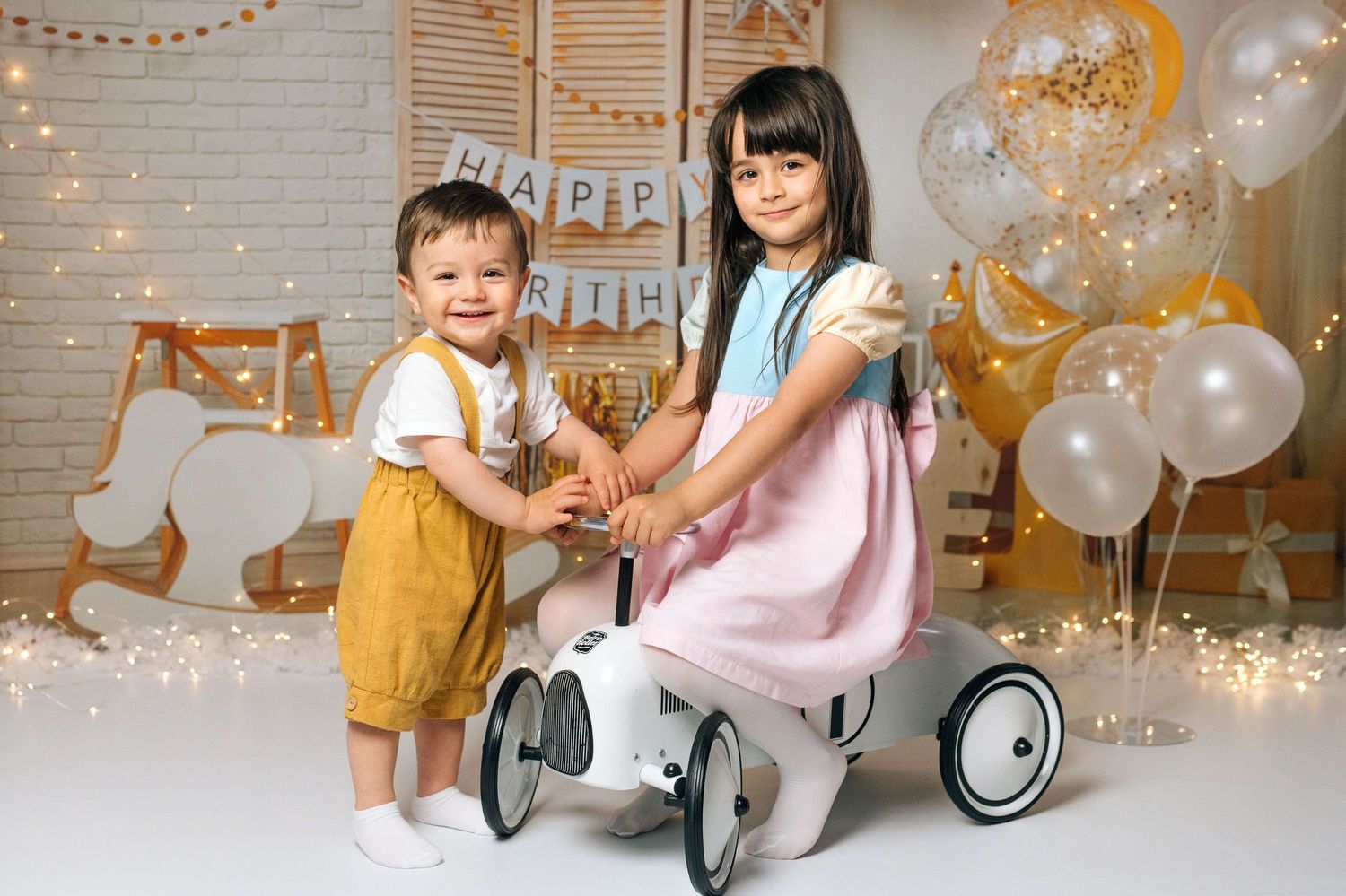 Two young children smile beside a toy car at a birthday party with balloons and festive decorations.