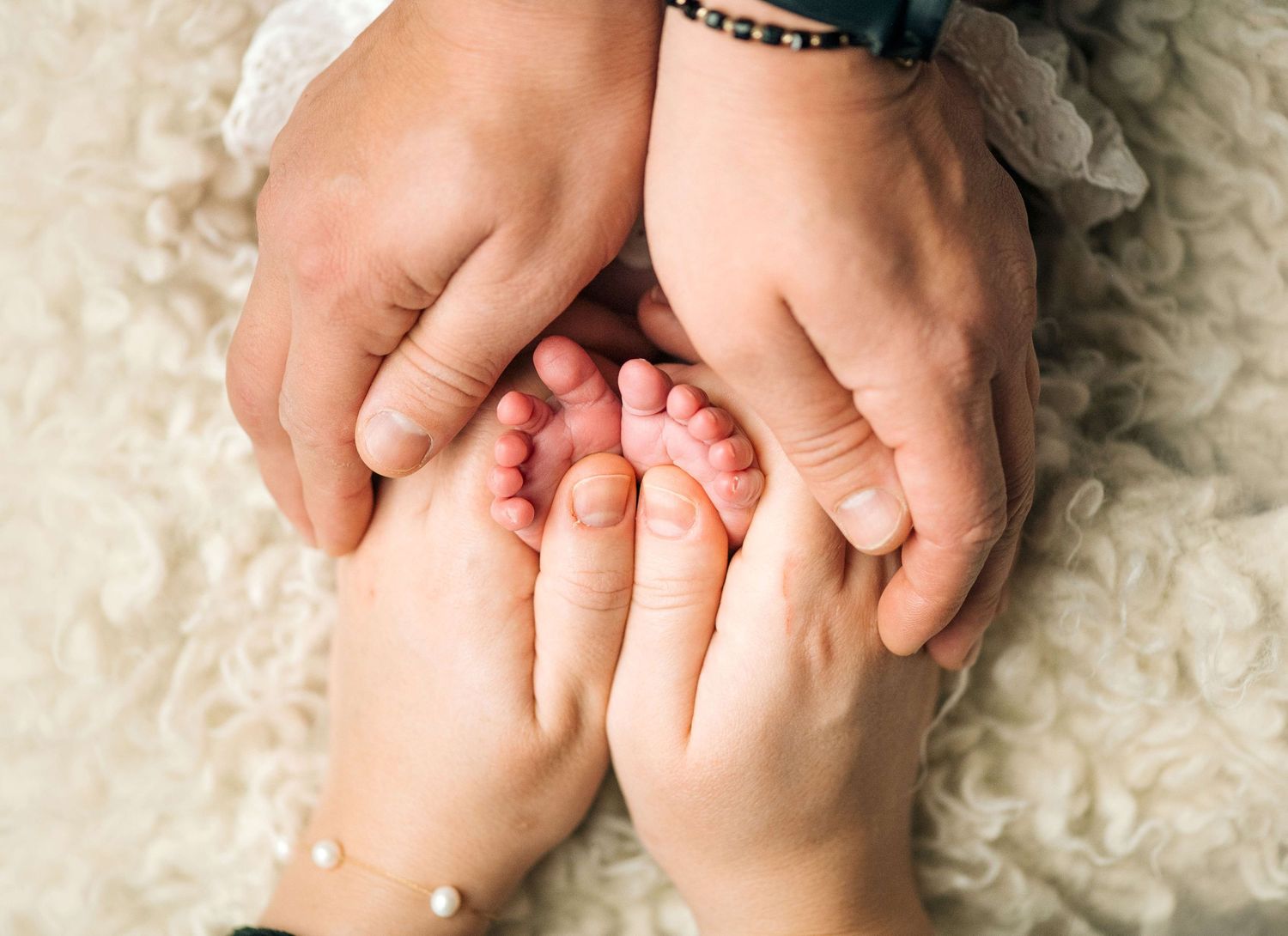 Adult hands gently cradling tiny newborn baby feet on a soft white blanket, a tender family moment.