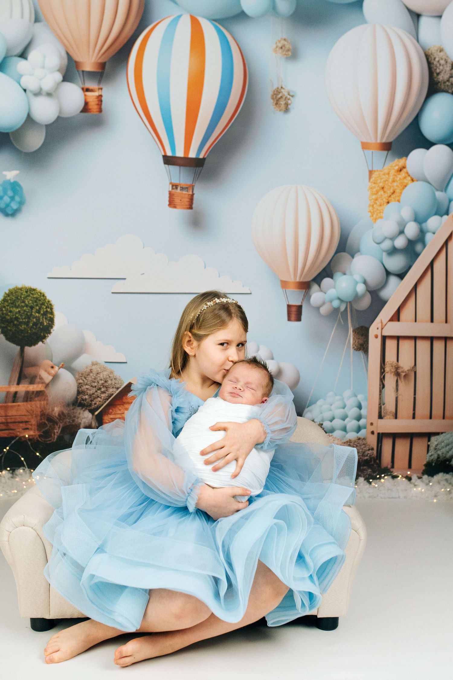 Young girl in blue tulle dress holds sleeping newborn baby in decorated studio with hot air balloon theme.