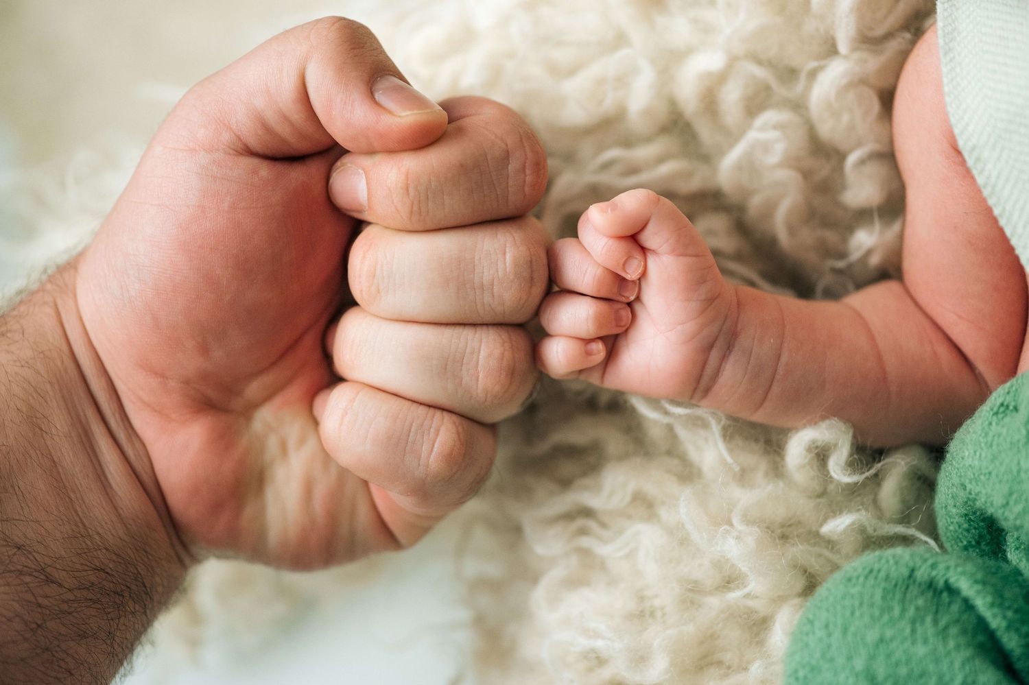 A father's fist bump with his newborn baby's tiny hand on a soft white blanket.