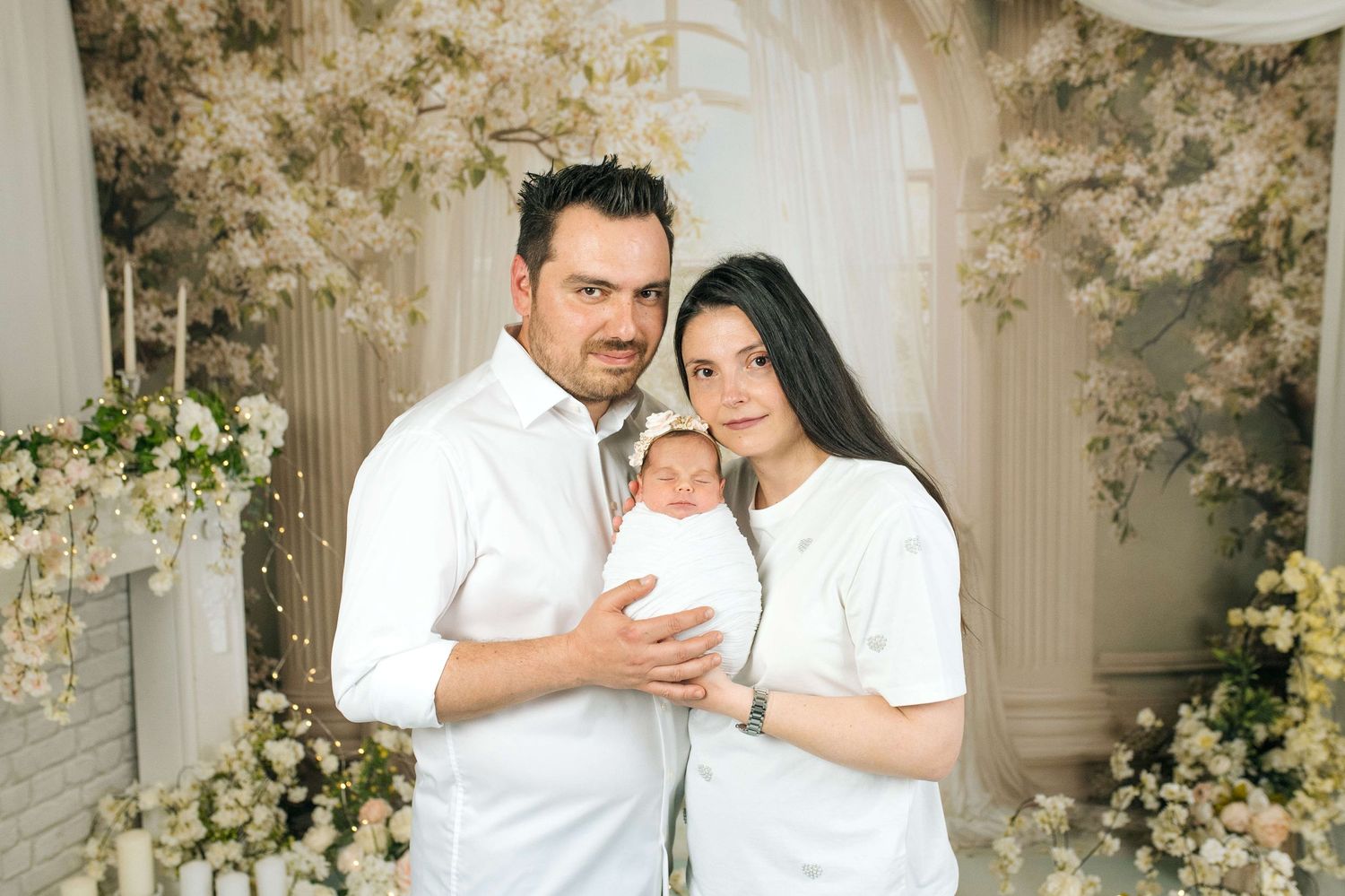 A couple in white outfits holds their newborn baby in a floral studio setting with white flowers and elegant decor.