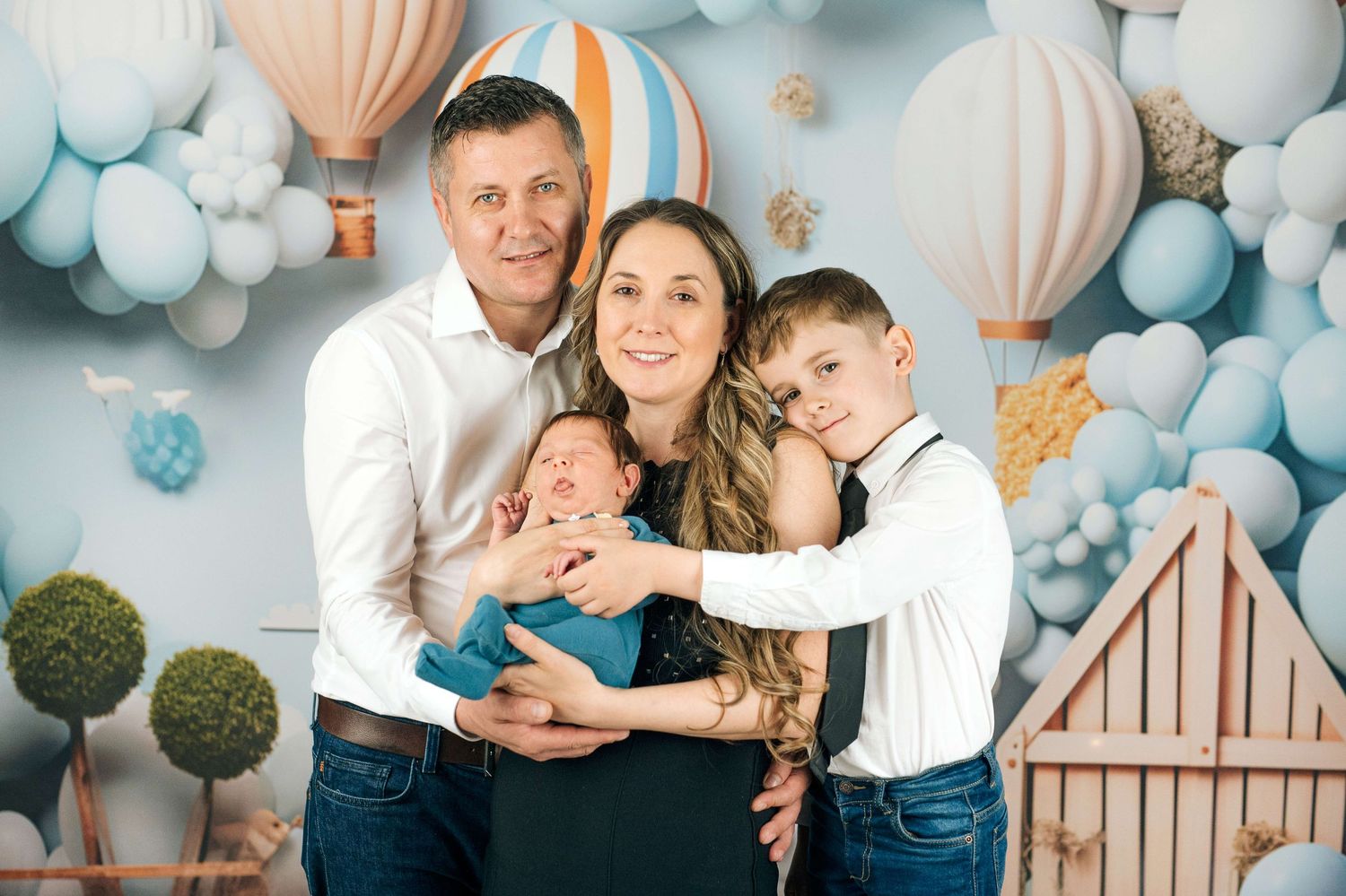 Family of four posing for a newborn photo shoot with hot air balloon and pastel balloon backdrop.