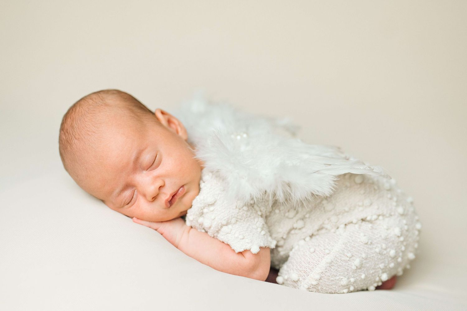 Peaceful newborn baby sleeping curled up with white fluffy angel wings wrap on a soft cream background.