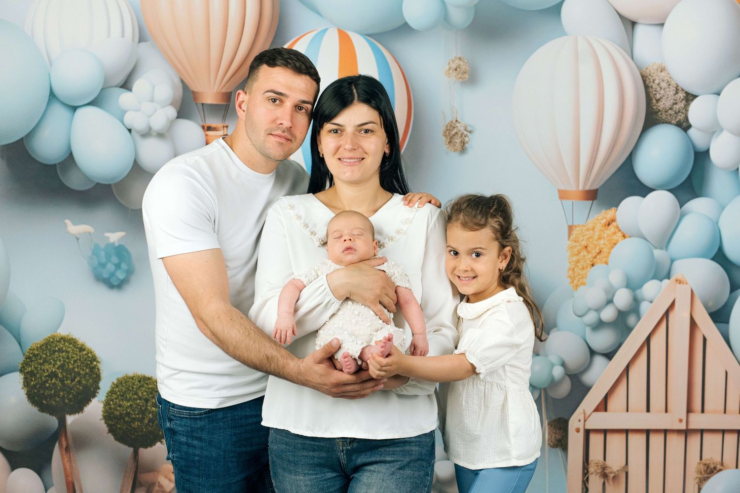 Family of four posing in front of a balloon and hot air balloon backdrop, parents holding newborn baby with young daughter.