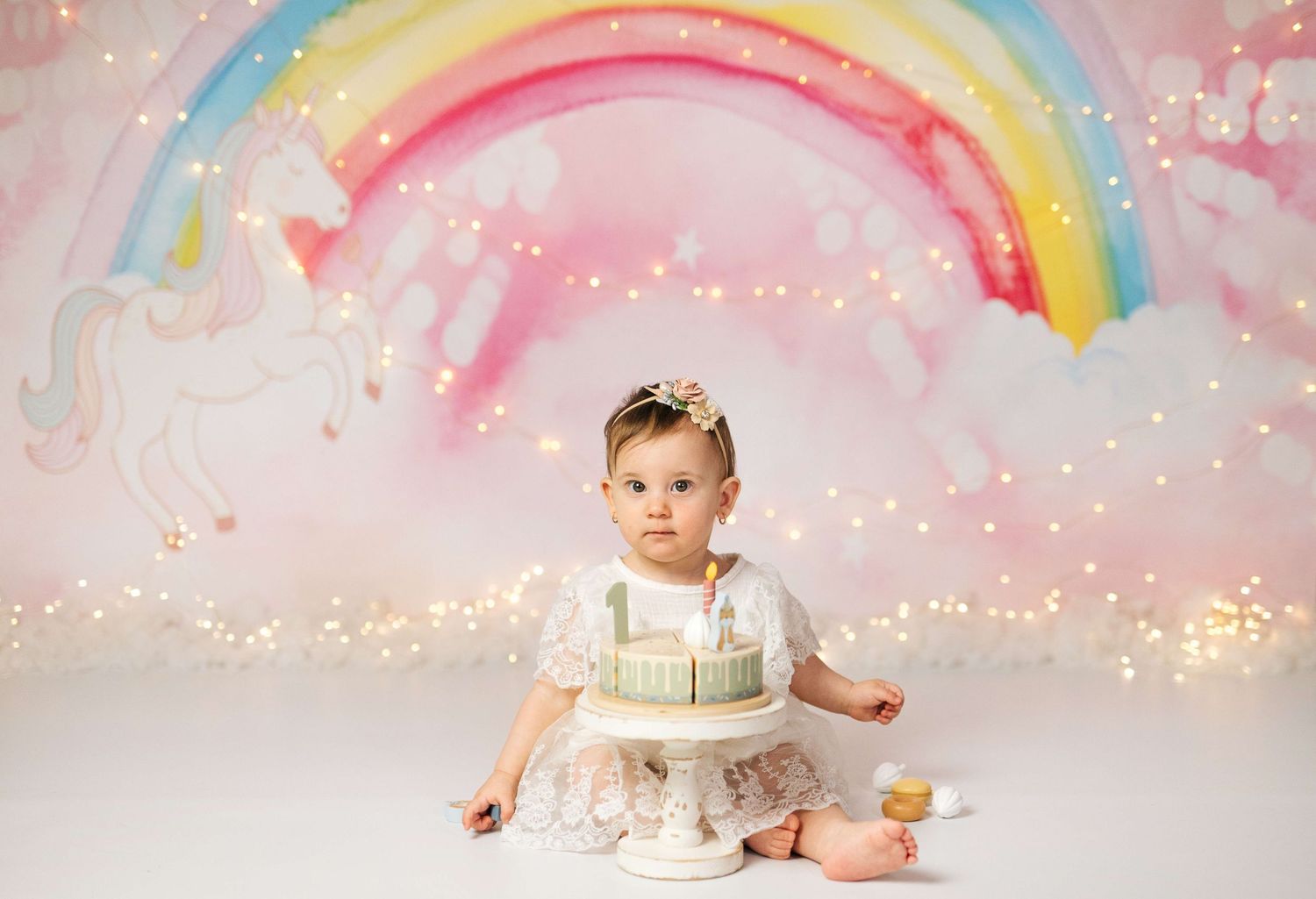 Baby girl in white dress sits before a 1st birthday cake with unicorn rainbow bokeh backdrop.