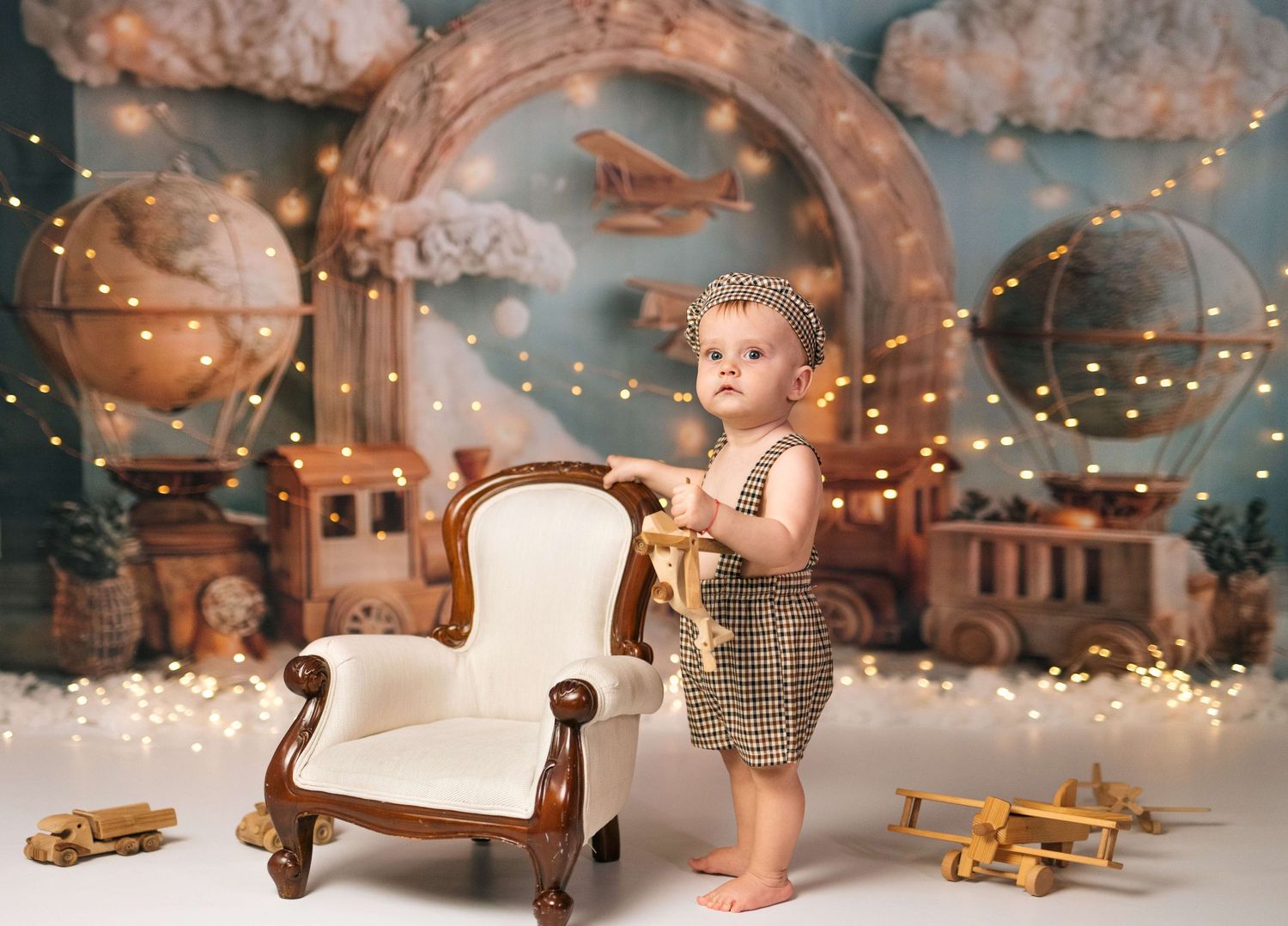 Baby in romper stands by vintage chair with wooden toy planes and fairy lights in a whimsical aviation-themed photo backdrop.