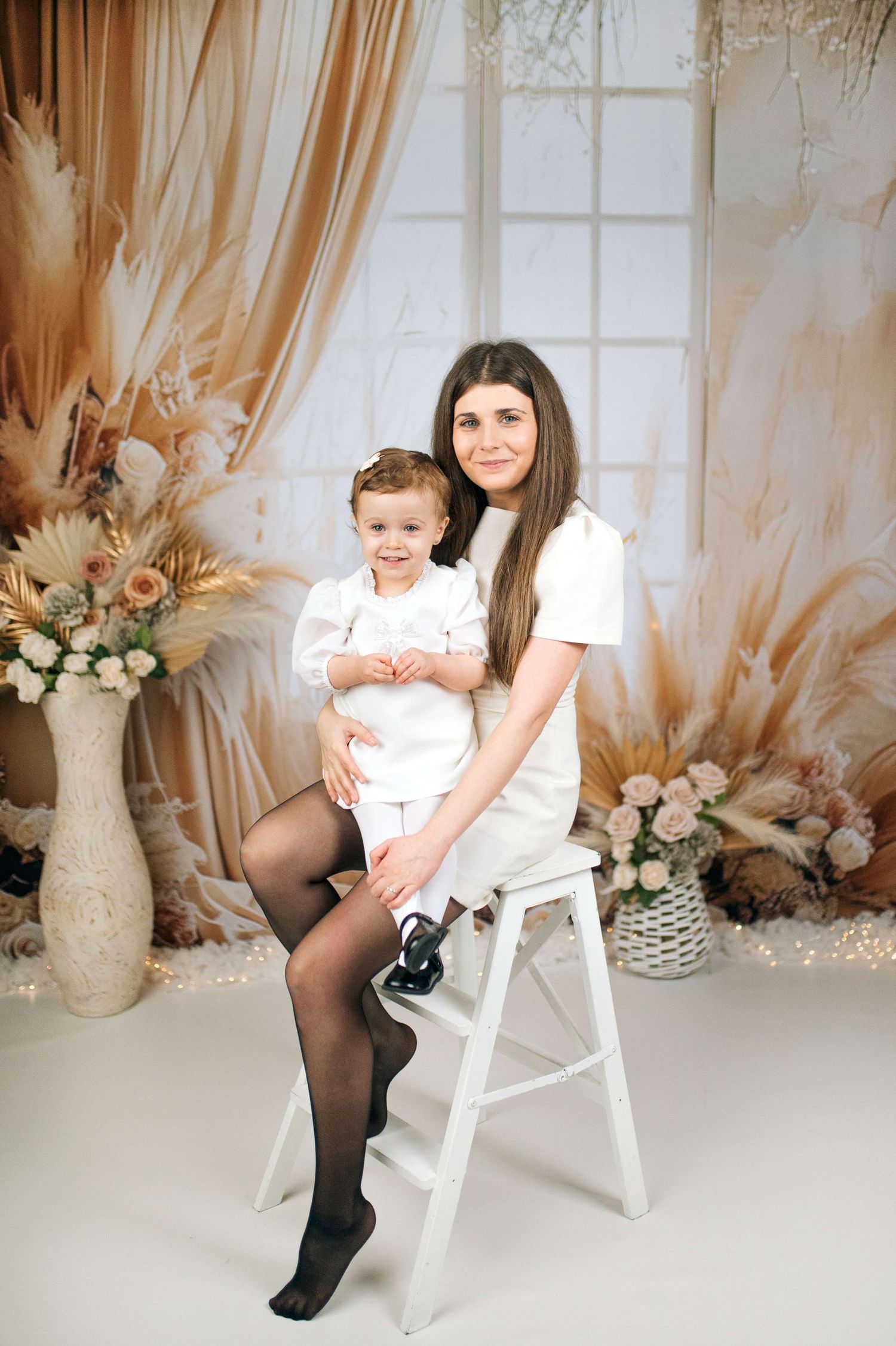 Young mother in white dress sits on stool holding toddler in white outfit, posing in boho floral studio backdrop.