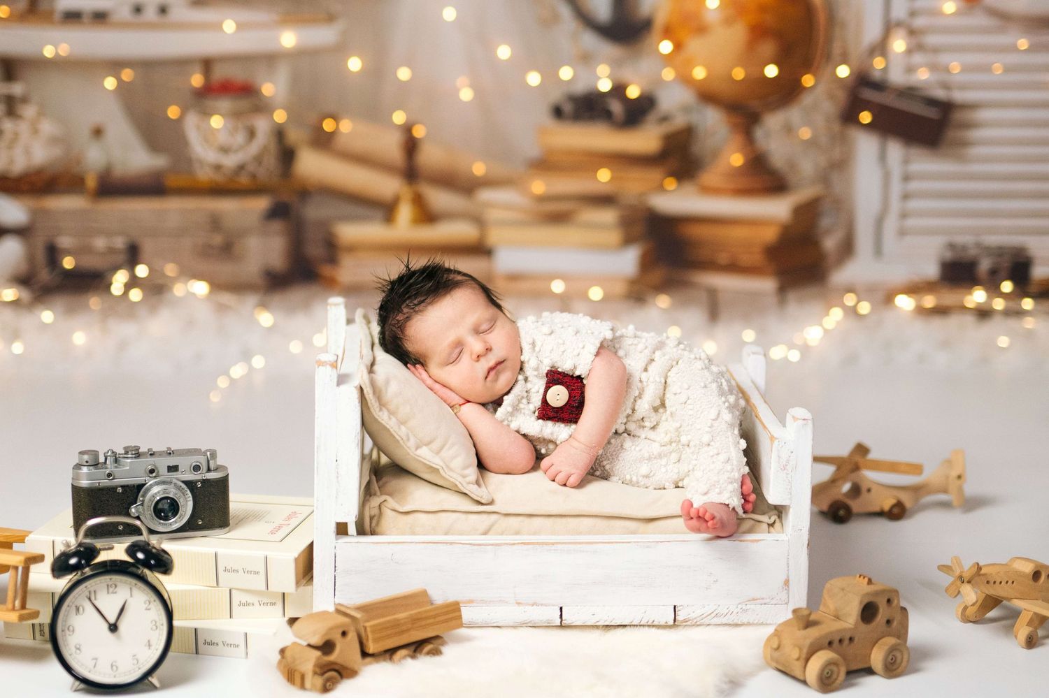 Newborn baby sleeping peacefully in a white miniature bed surrounded by wooden toys and fairy lights.