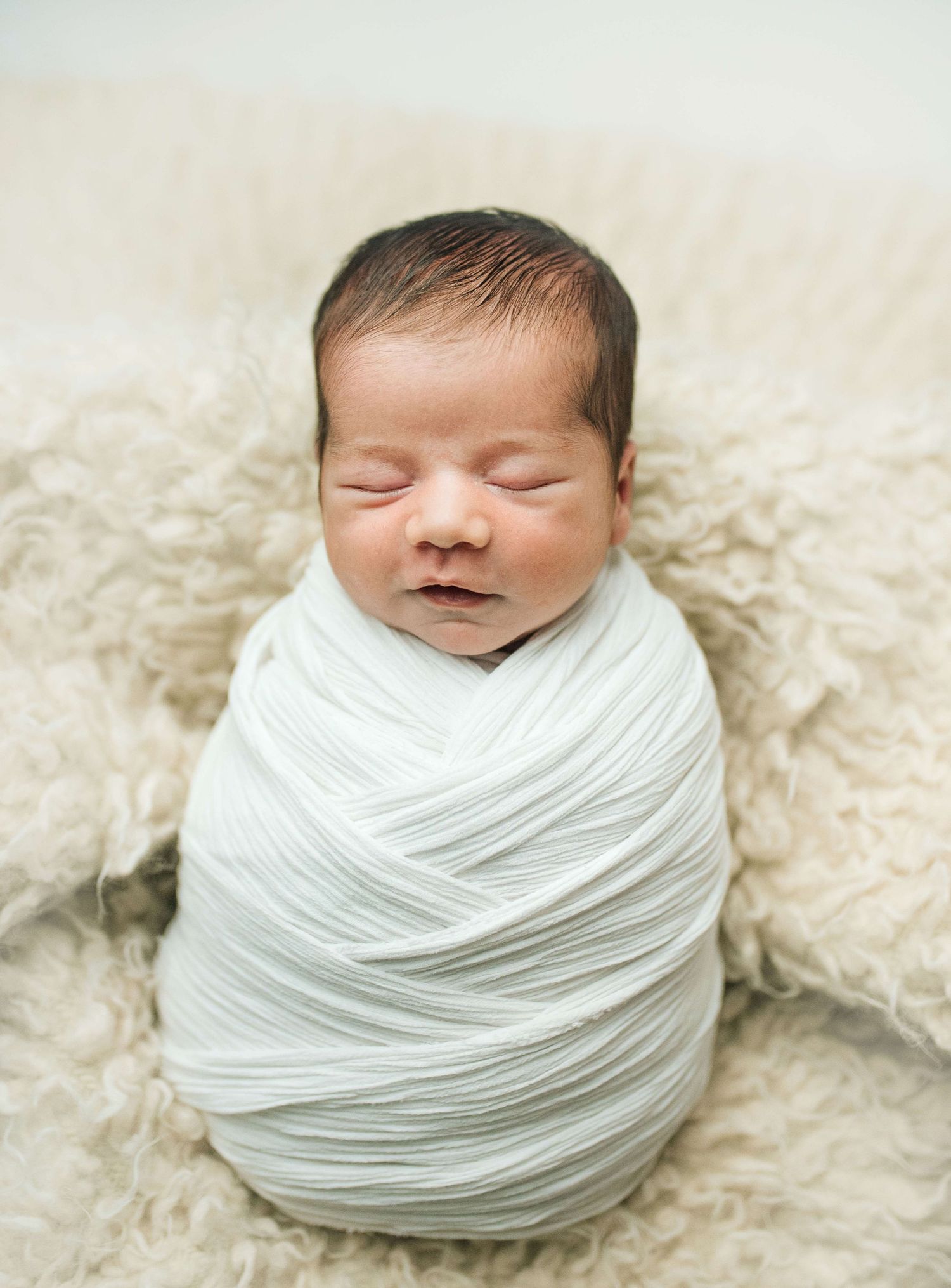 Newborn baby swaddled in white wrap sleeping peacefully on fluffy cream blanket in professional studio photo.