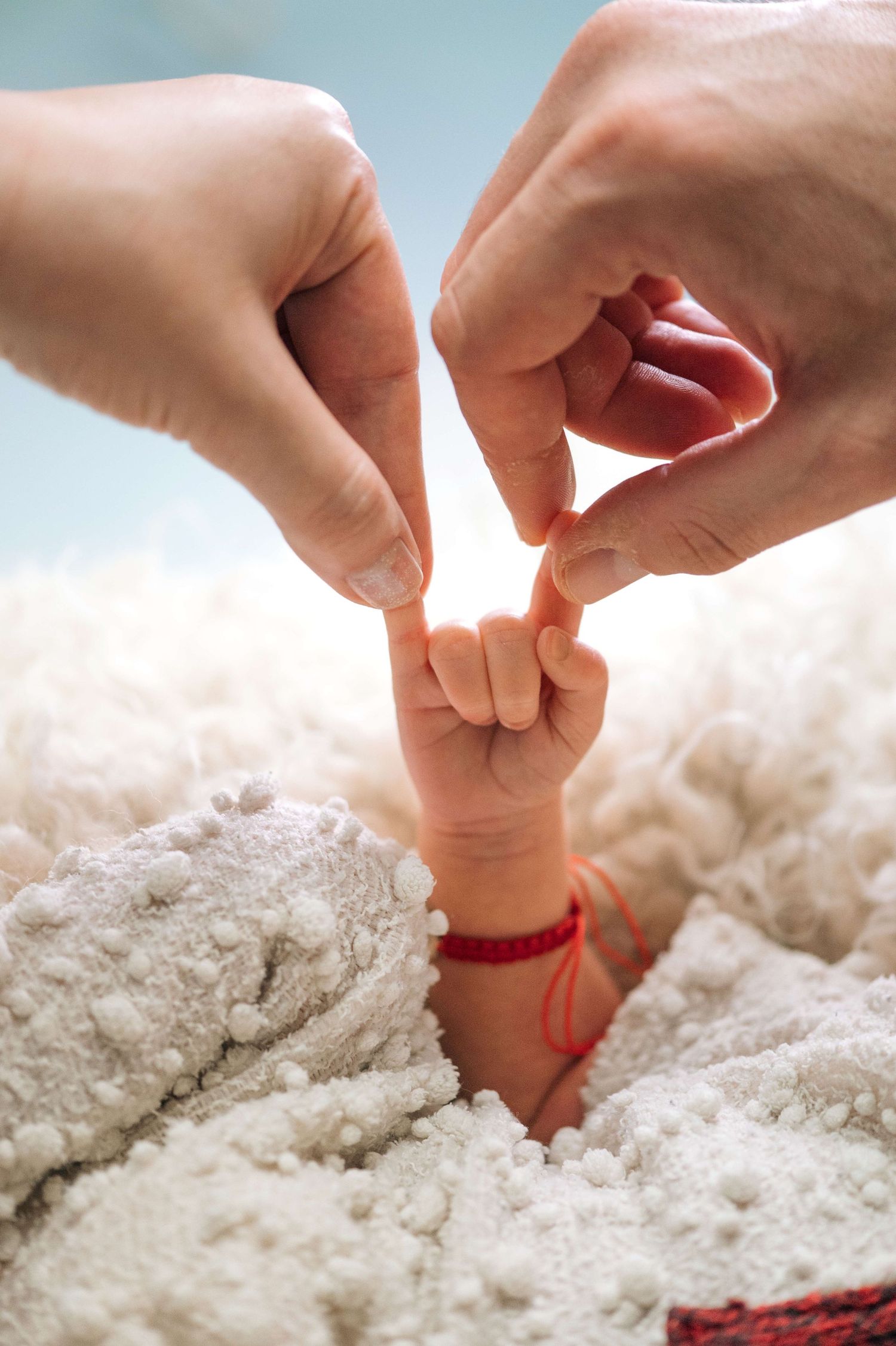 Newborn baby hand gripping parents fingers on white fluffy blanket, with red bracelet on tiny wrist.